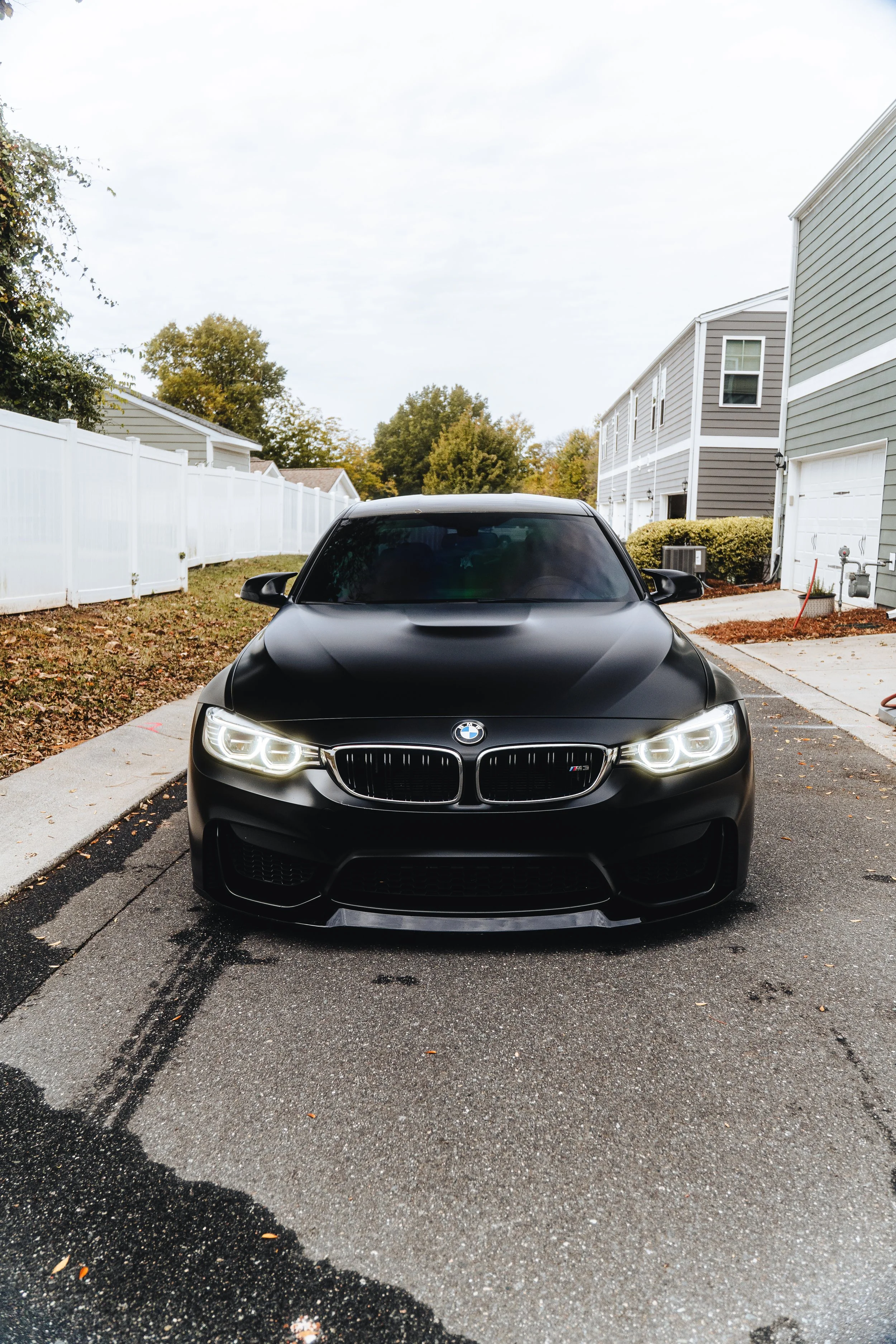 Black BMW car parked in a residential driveway with a white fence and houses in the background.
