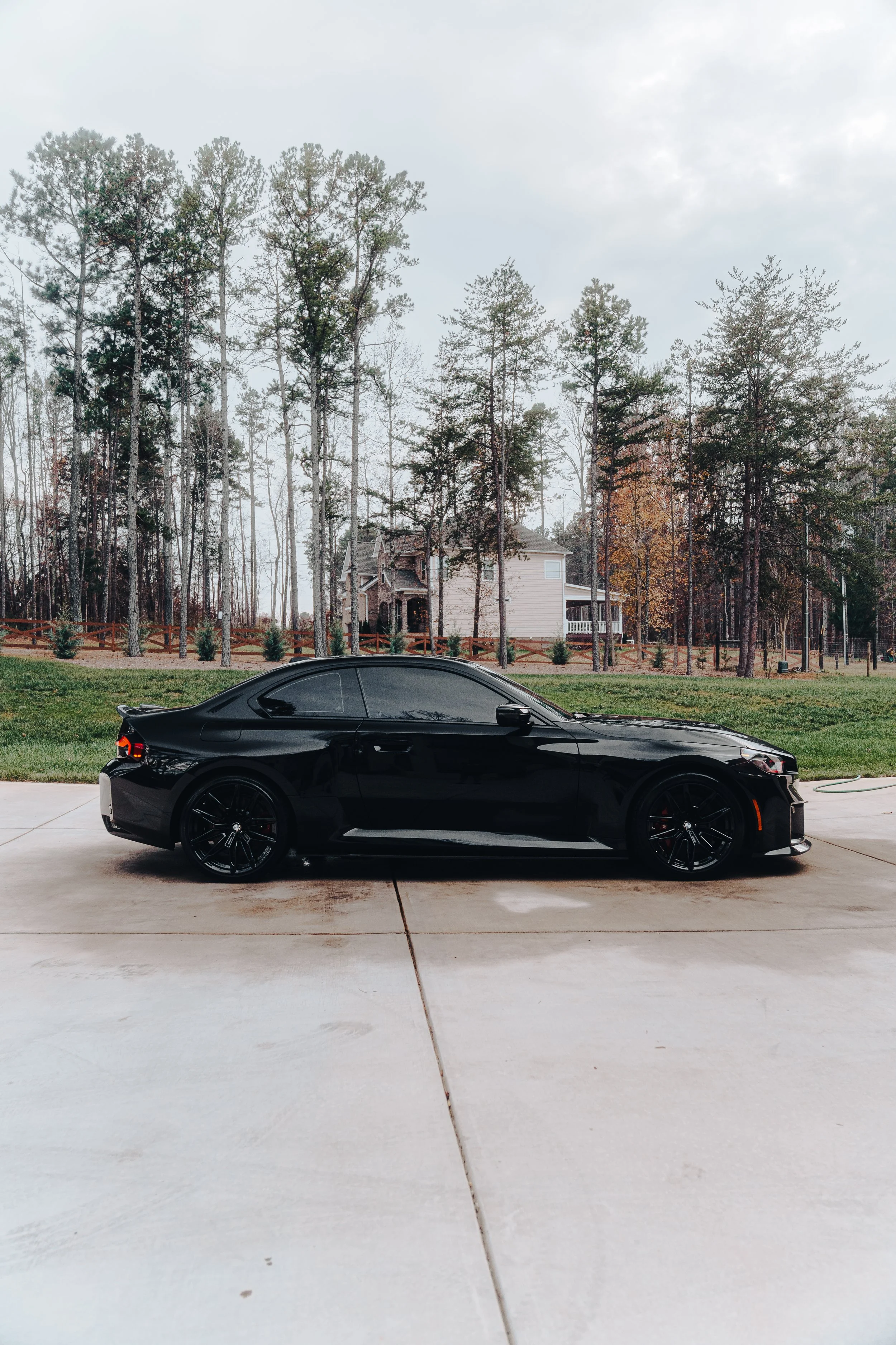 A sleek black sports car parked on a concrete driveway with a suburban house and tall trees in the background.