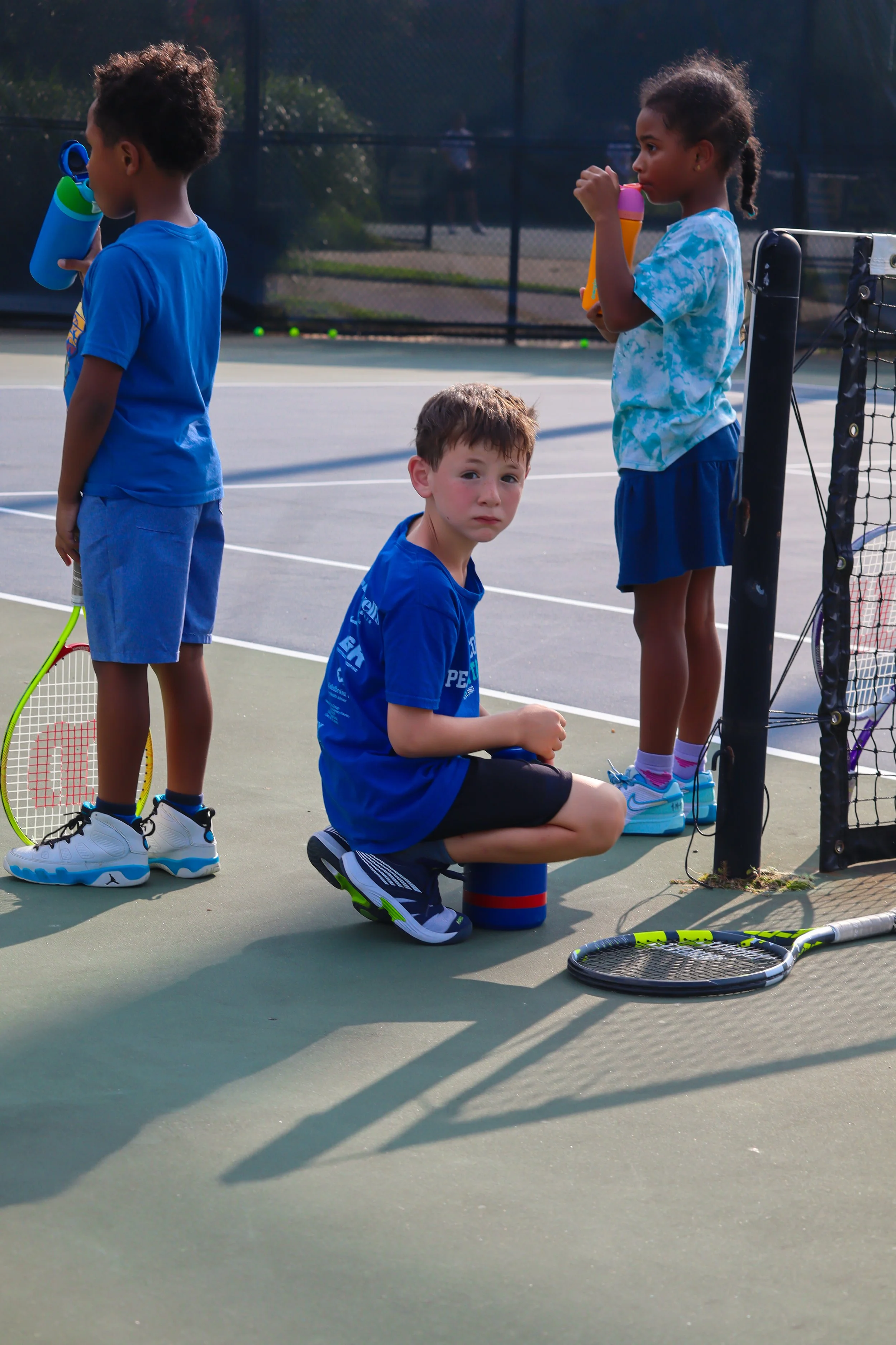 Three children on a tennis court, two girls drinking from water bottles and a boy sitting on a small stool, holding a tennis racket and looking at the camera.