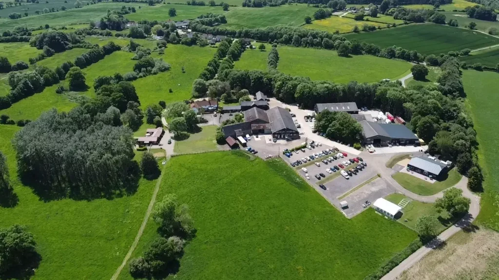 Aerial view of a rural farm with buildings, parking lot, and surrounding green fields and trees.