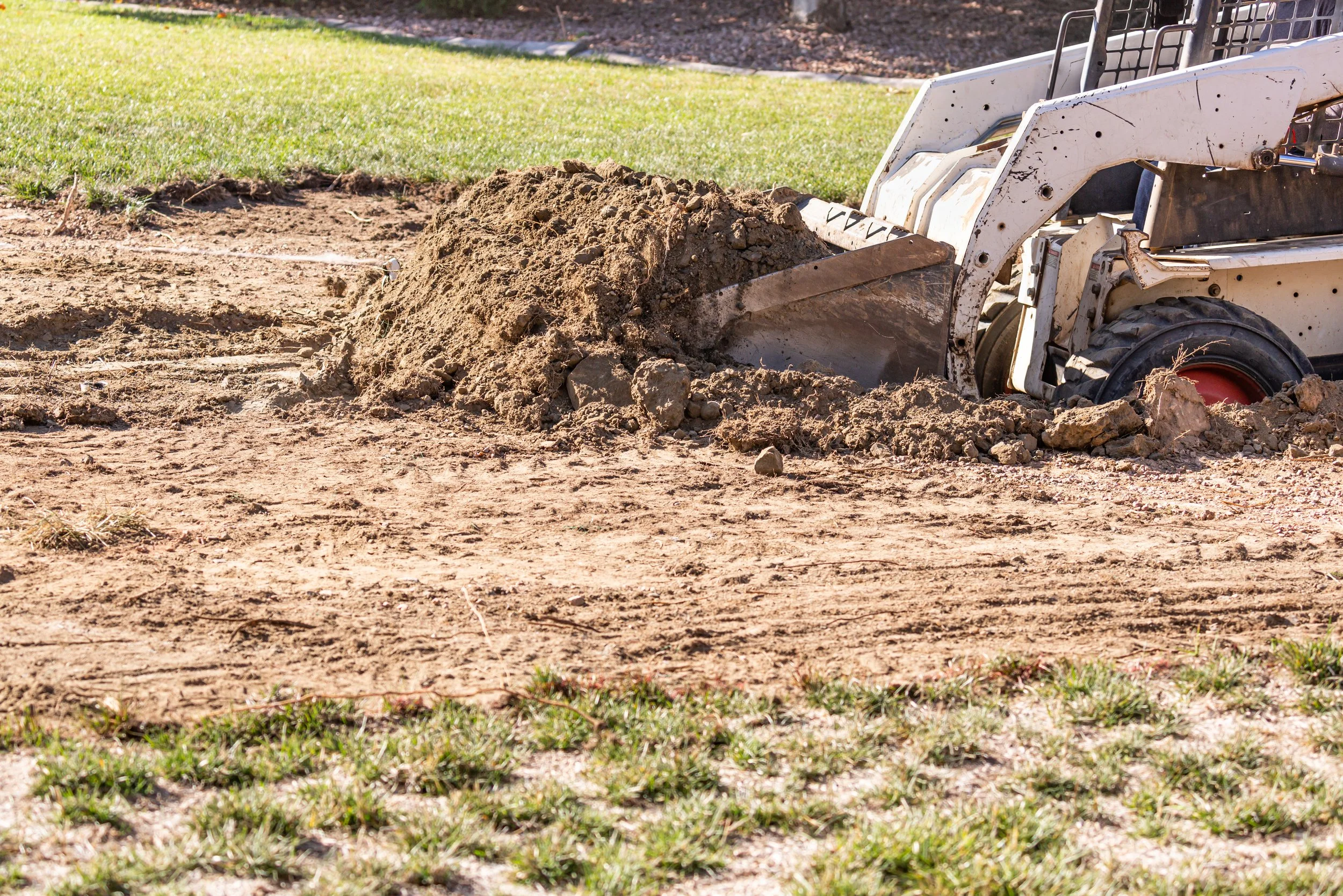 A small construction vehicle, like a skid steer loader, spreading dirt on a patch of ground with grass nearby.