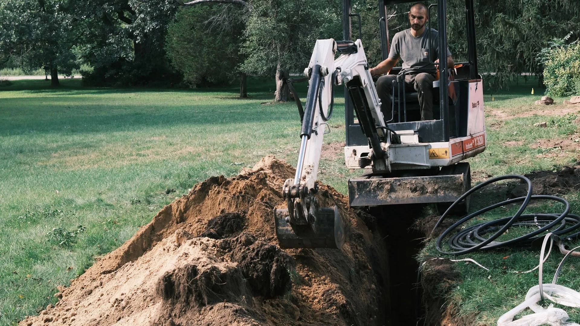 A man operating a small excavator digging a trench in a grassy area with trees in the background.