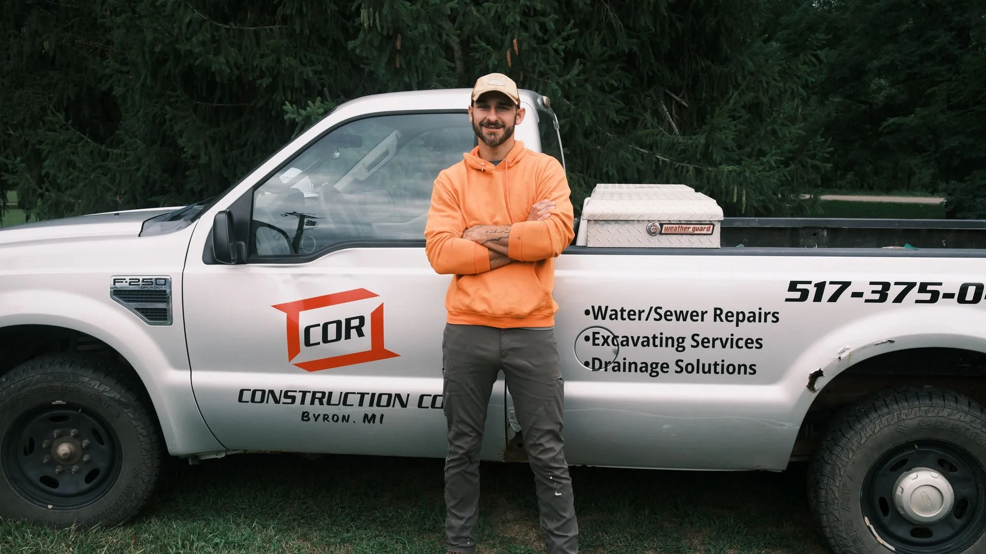 A man standing with arms crossed in front of a white construction service pickup truck on a grassy area, with a background of trees. The truck has Text advertising water and sewer repairs, excavating services, and drainage solutions, and includes a phone number. The man wears a beige cap, orange hoodie, and gray pants, and is smiling.