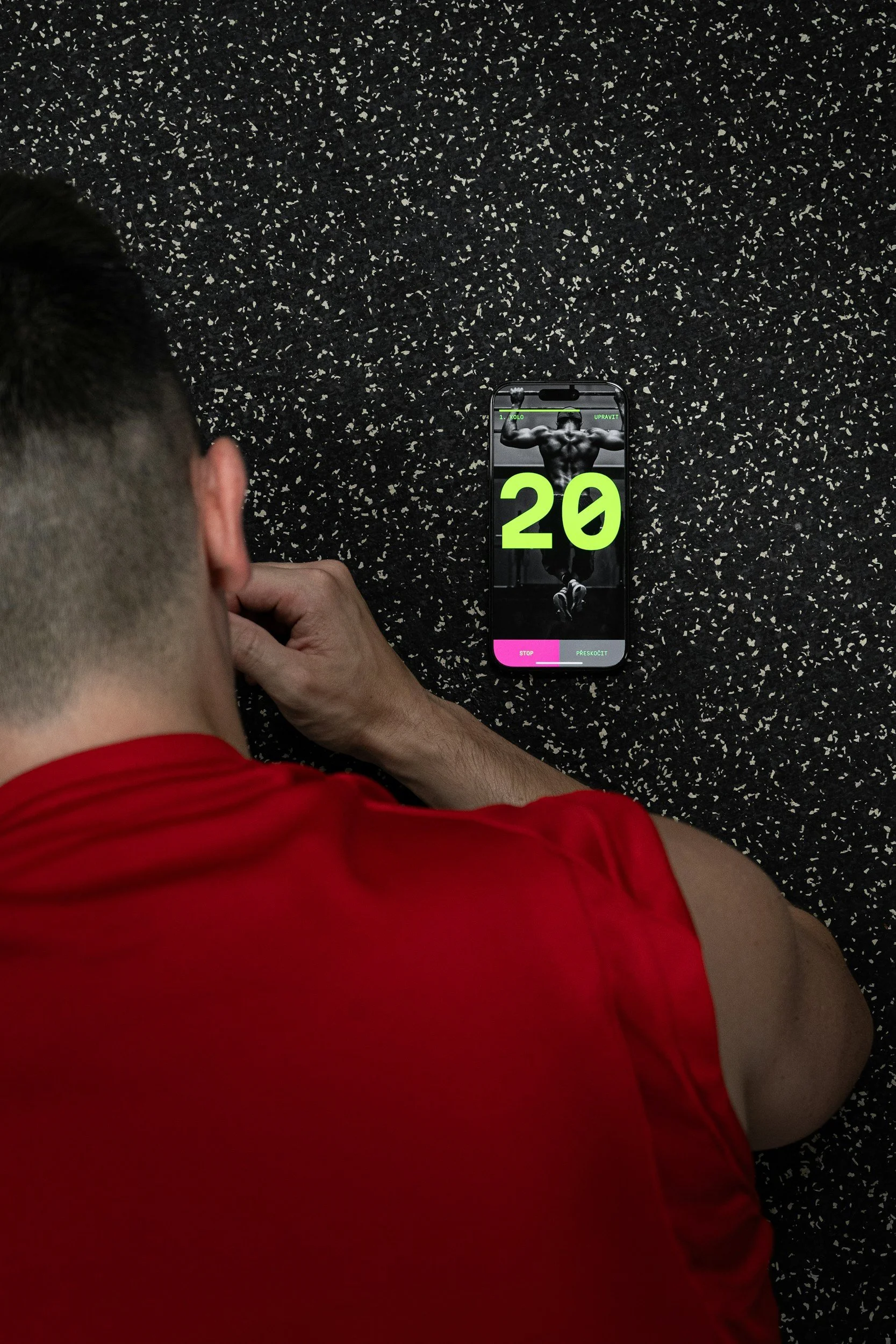 A person wearing a red shirt with short black hair is lying on a black speckled exercise mat. They are looking at a smartphone displaying an image of a muscular man flexing his back muscles with the number 20 prominently shown on the screen.