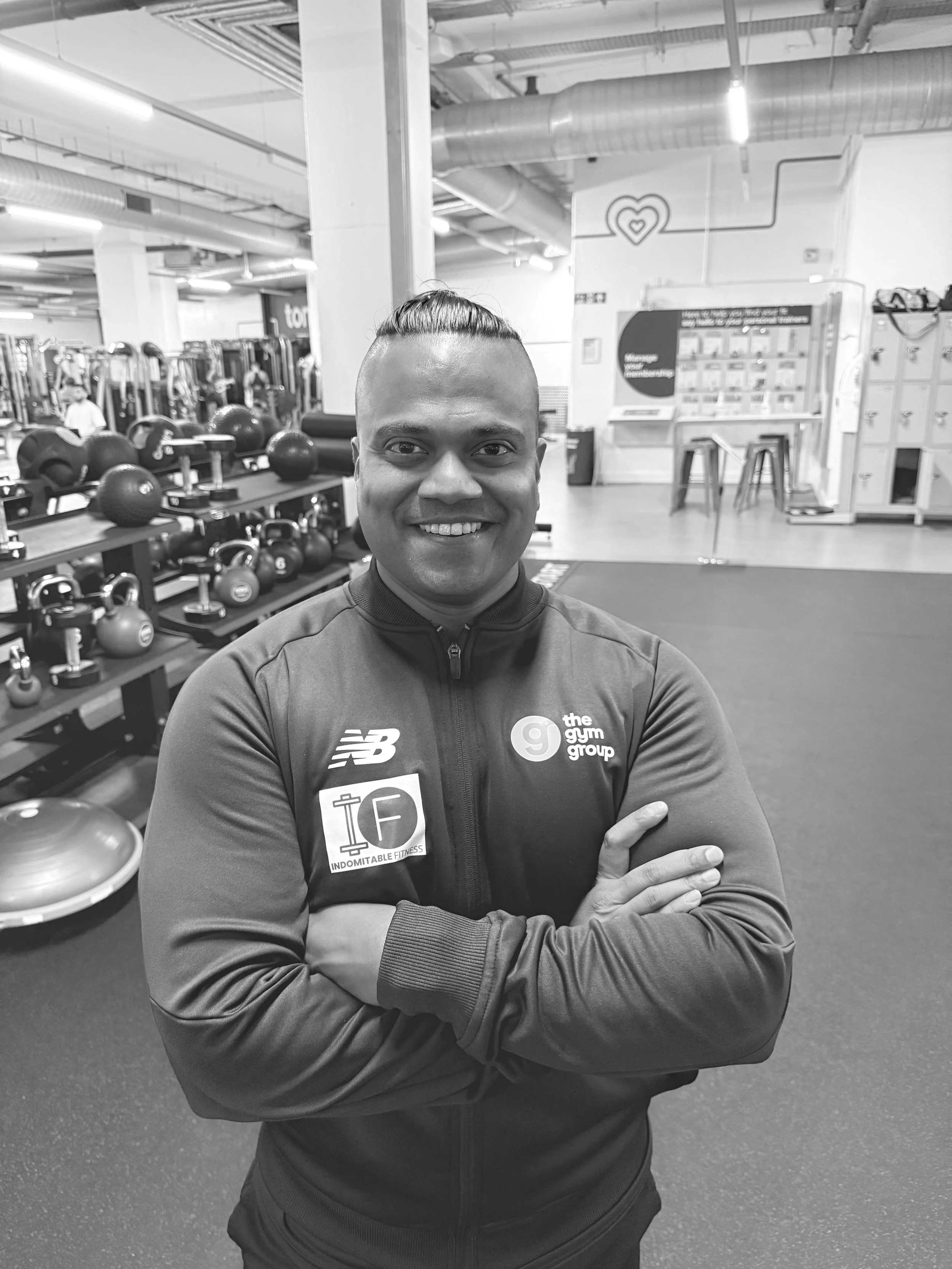 A smiling man with a samurai hairstyle taking a selfie in a gym, with gym equipment including kettlebells and exercise balls visible in the background.