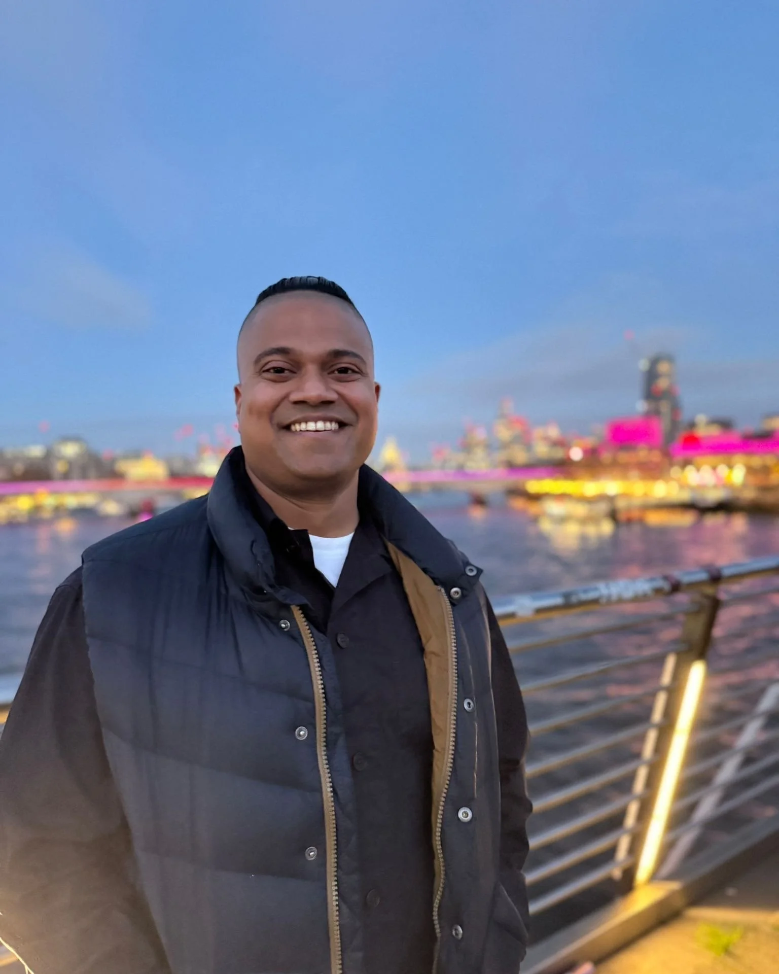 A smiling man standing outdoors with a city skyline in the background, illuminated at dusk or night, near a waterfront with a railing.