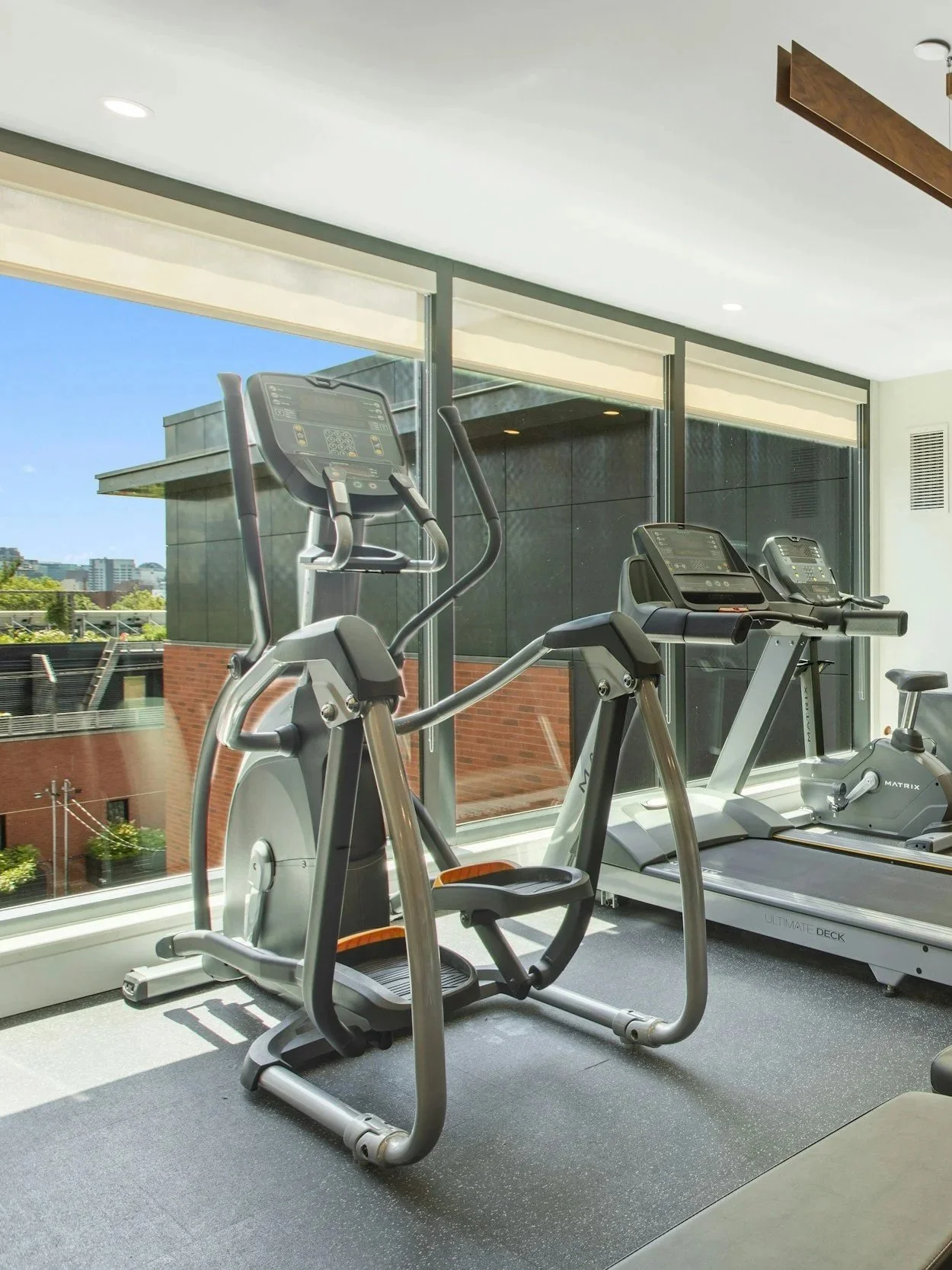 A gym room with two exercise machines in front of large windows showing a cityscape and a building. The gym has a bright, modern design with a white ceiling and grey floor.