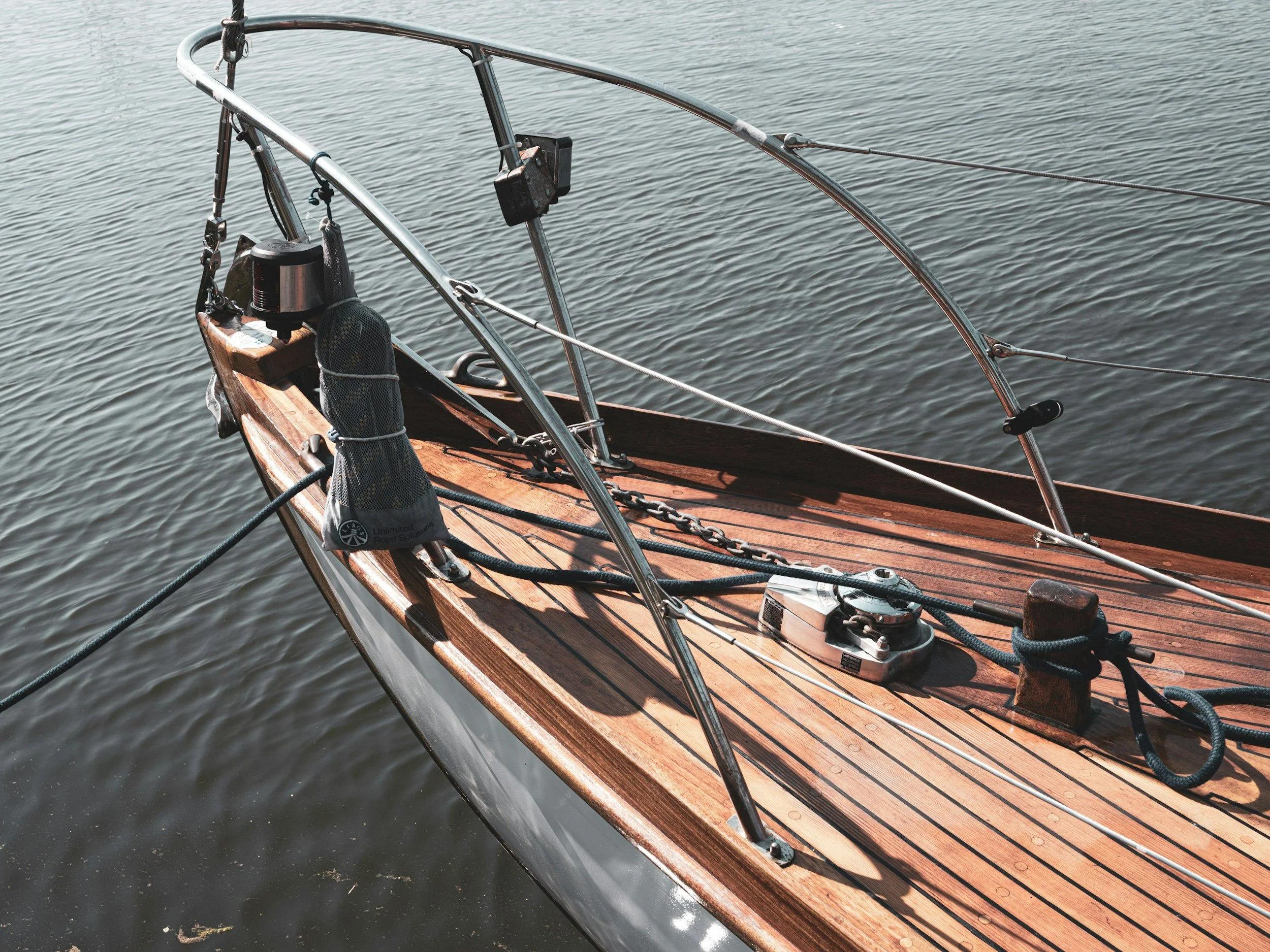 Close-up of a wooden sailboat deck with metal railings, ropes, winch, and hardware, floating on calm water.