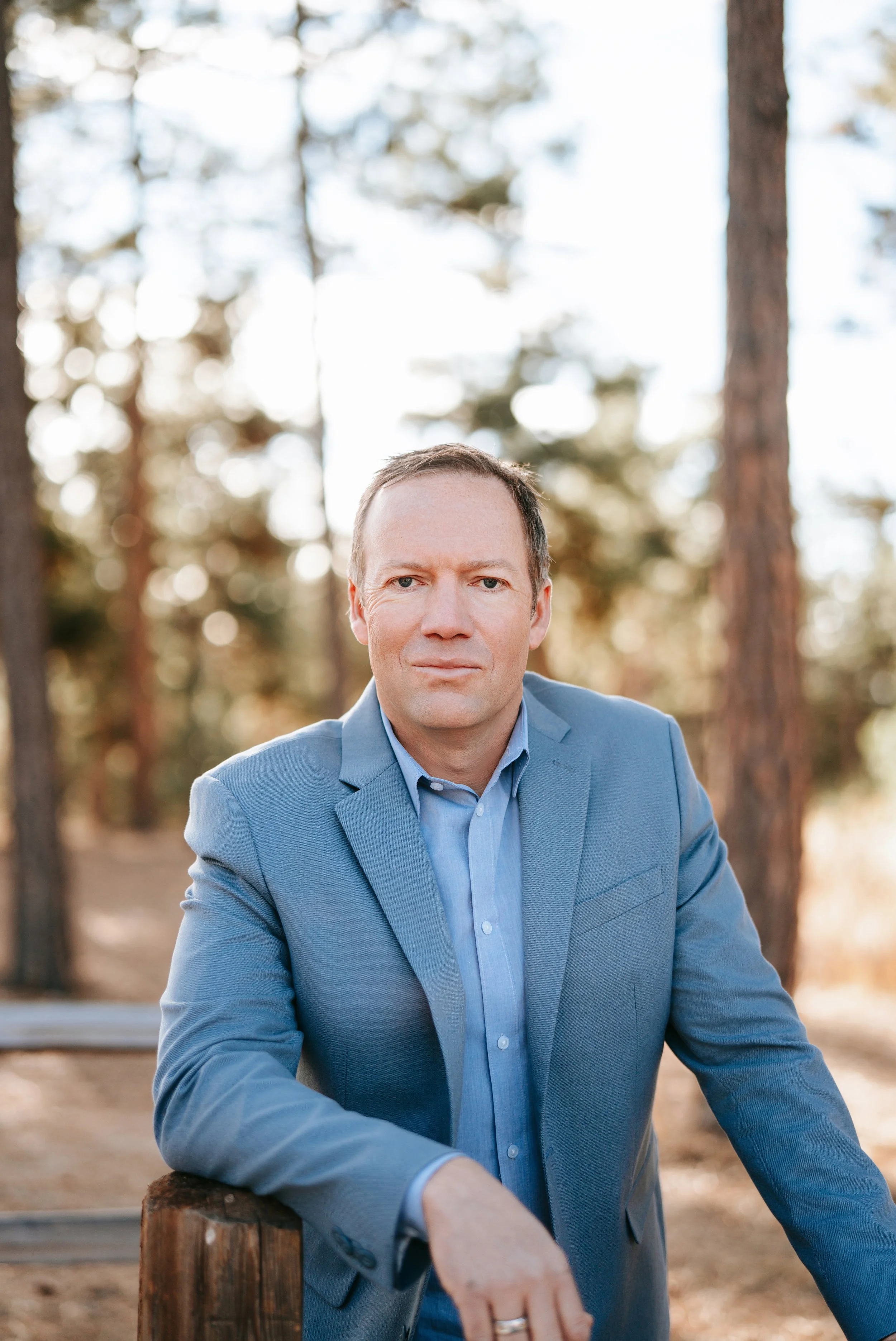 A man in a light blue suit jacket and shirt sitting outdoors with trees in the background.