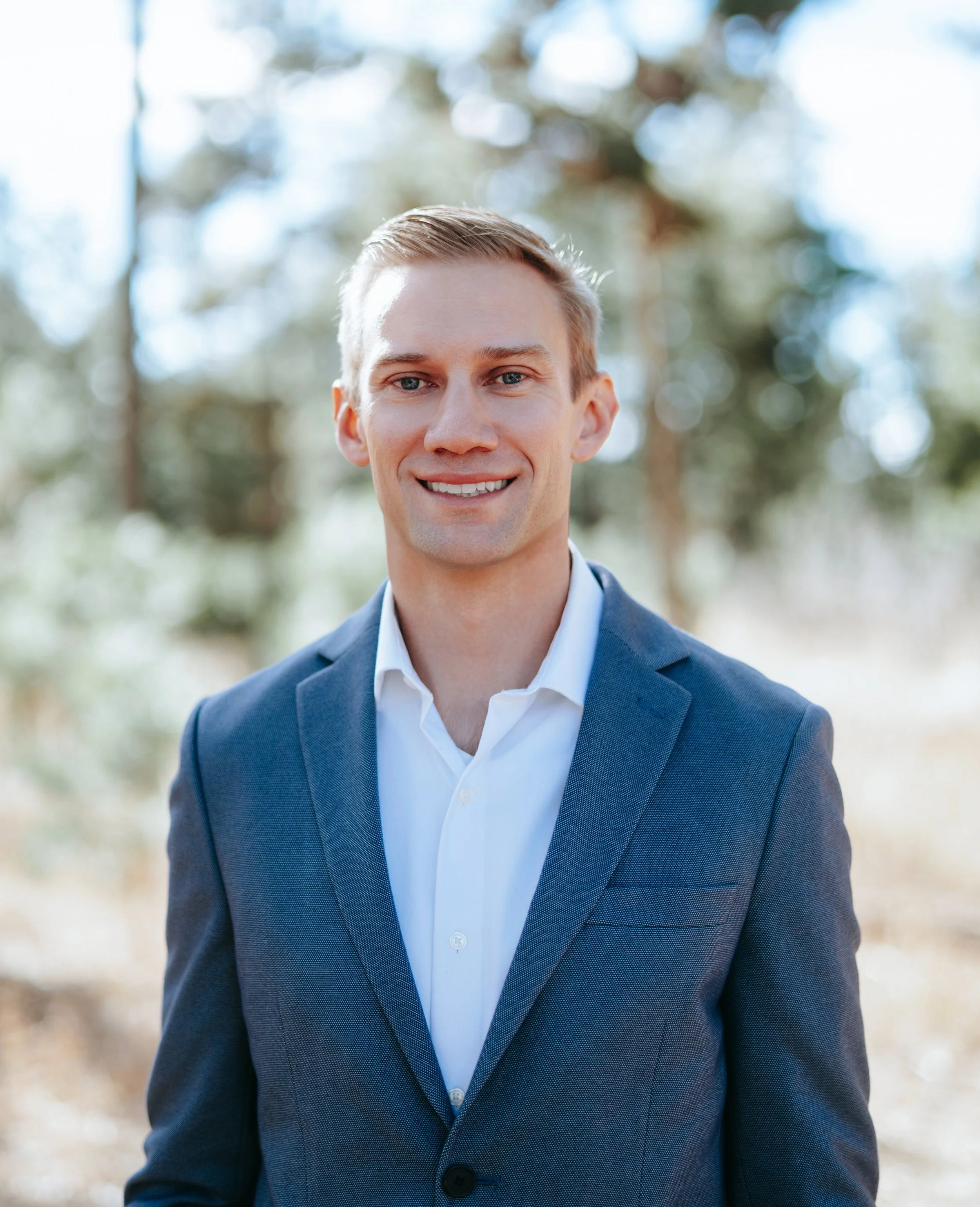 A young man in a navy blazer and white shirt standing outdoors with trees in the background, smiling at the camera.