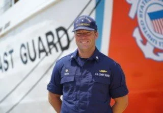 A Coast Guard officer in uniform standing in front of a boat with 'Coast Guard' text and a large orange and white structure in the background.