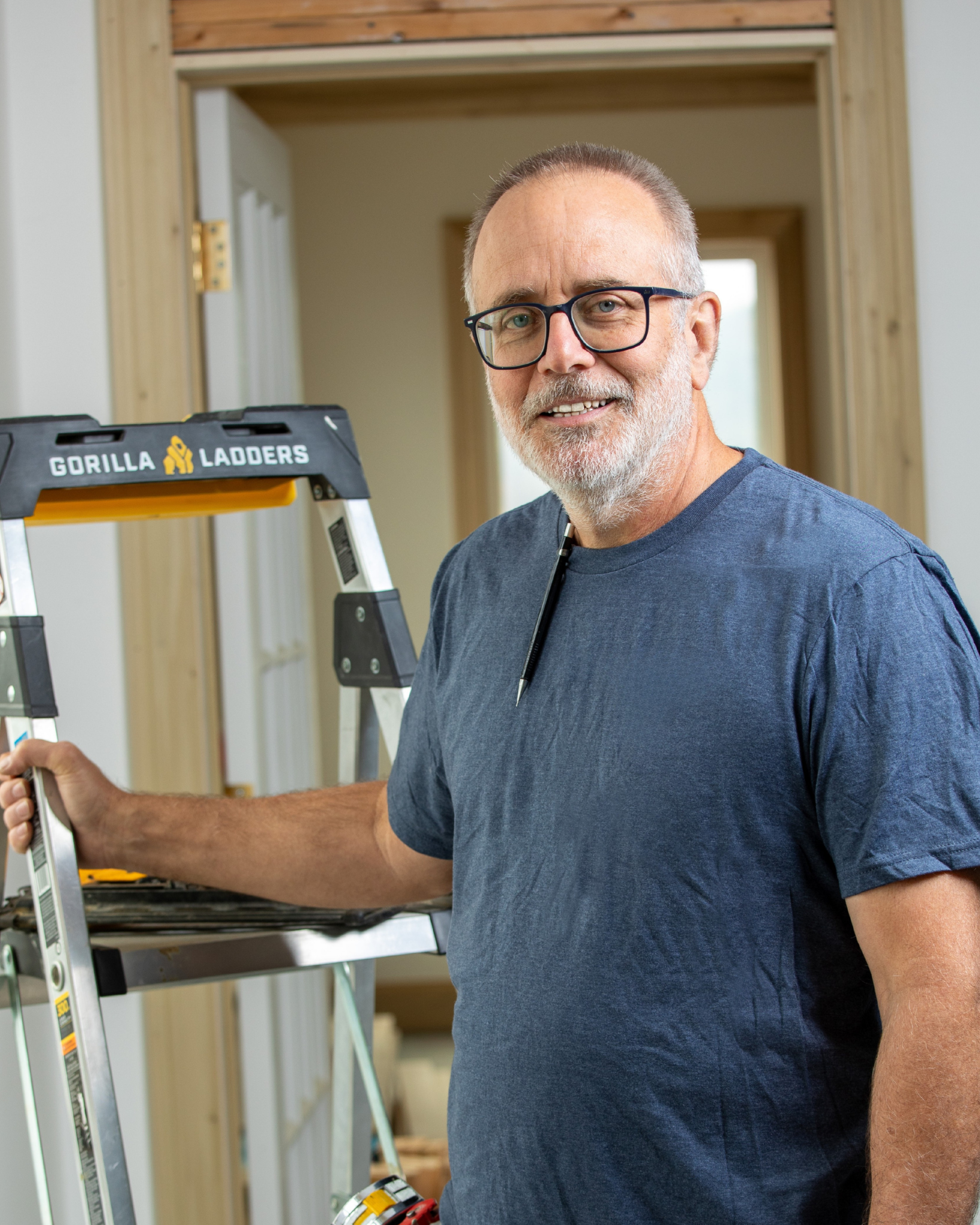 Bart Yeary is a smiling older man with glasses and a white beard standing indoors beside a step ladder, holding onto the ladder.
