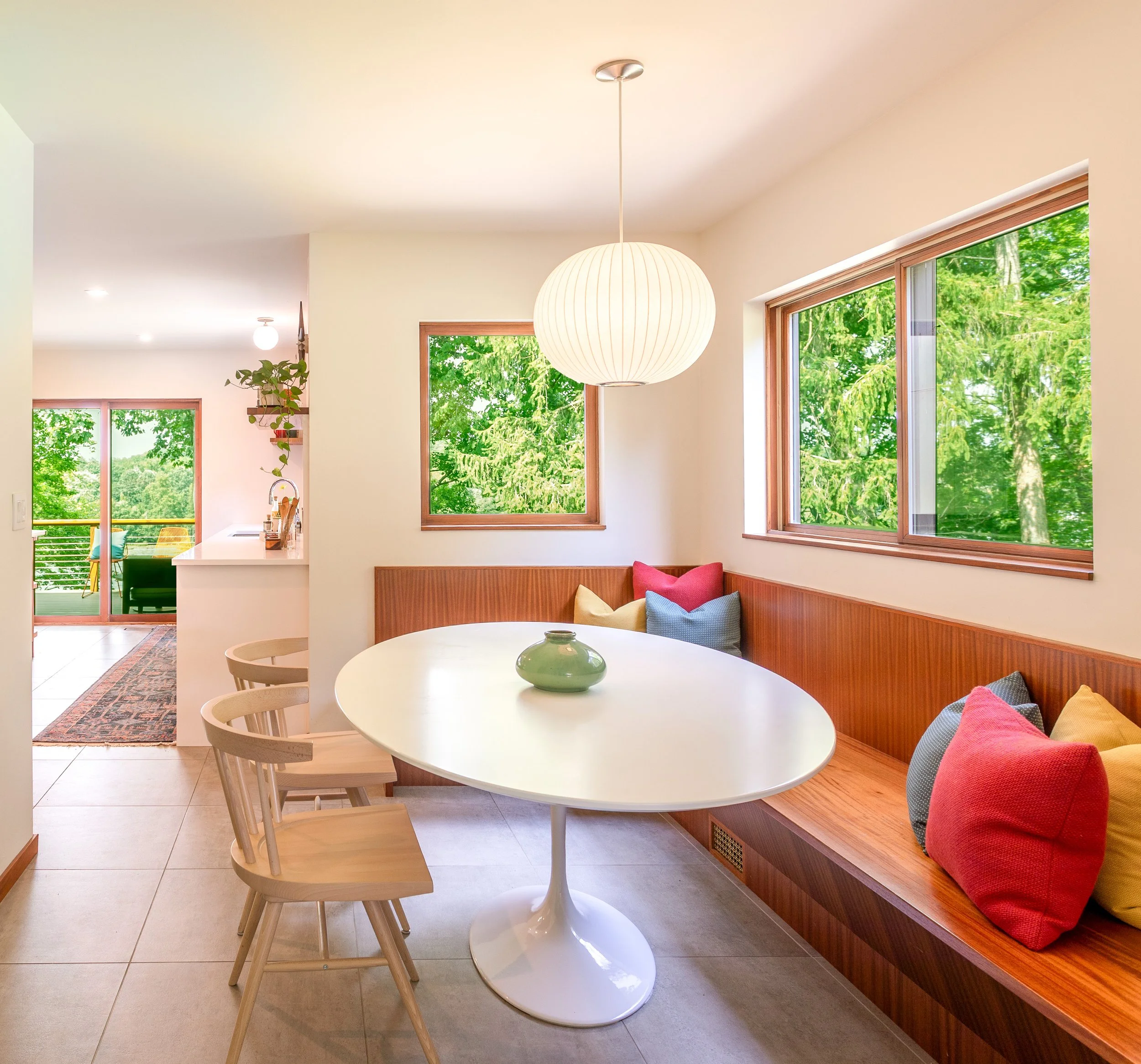 A bright kitchen and dining area with large windows showing green trees outside, a white round table with a green vase, wooden chairs, a built-in wooden bench with colorful pillows, and a white paper lantern light fixture.