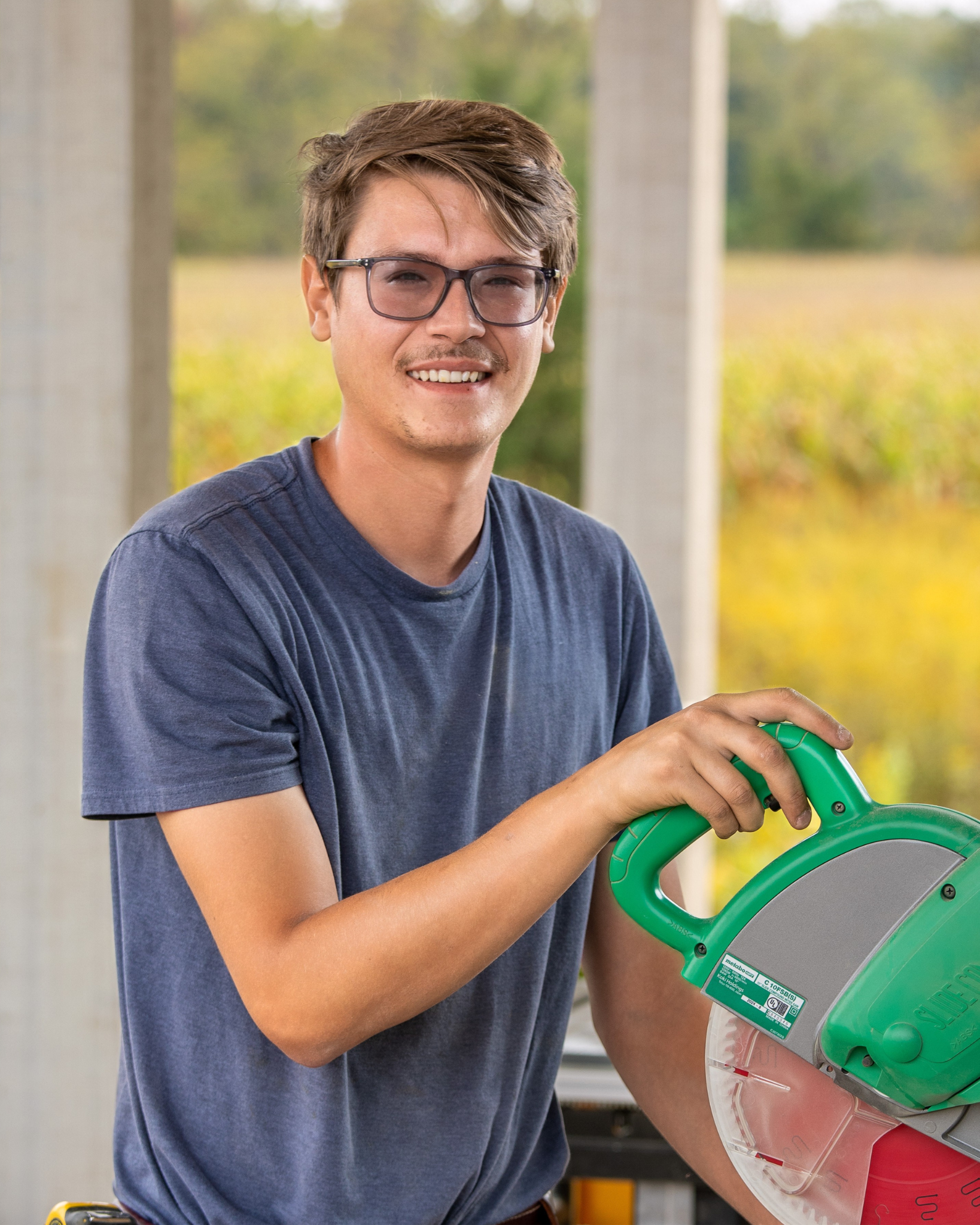 Gabriel Hardy is a young man with glasses and a blue t-shirt is smiling and holding a green and gray power saw for cutting wood or material.