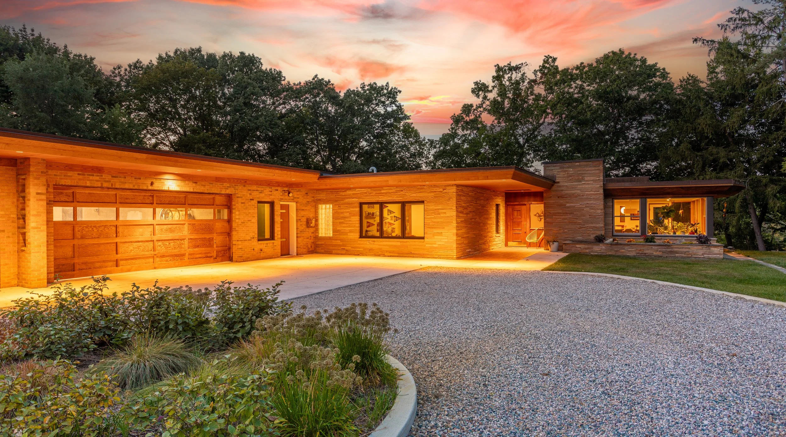 Mid-century modern house with brick and stone exterior, surrounded by a gravel driveway and landscaped garden, illuminated at dusk. This historic home was designed and built by Architect Ralph Stephens Gerganoff (R.S. Gerganoff).