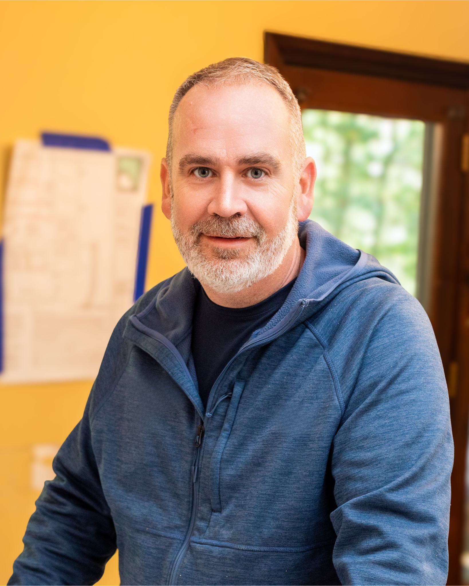 Jason Pace is a middle-aged man with a gray beard and short hair, smiling, wearing a blue zip-up hoodie, standing indoors near a window with green trees outside.