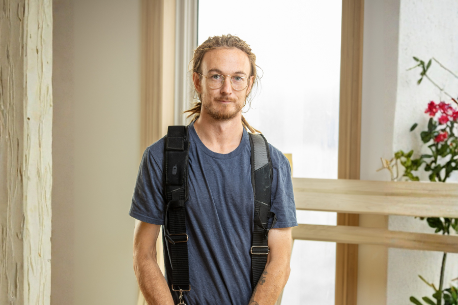 Josh Morris is a young man with long blond hair, glasses, and a beard, wearing a blue t-shirt and backpack, standing indoors near a window with a wooden railing and a plant with pink flowers nearby.