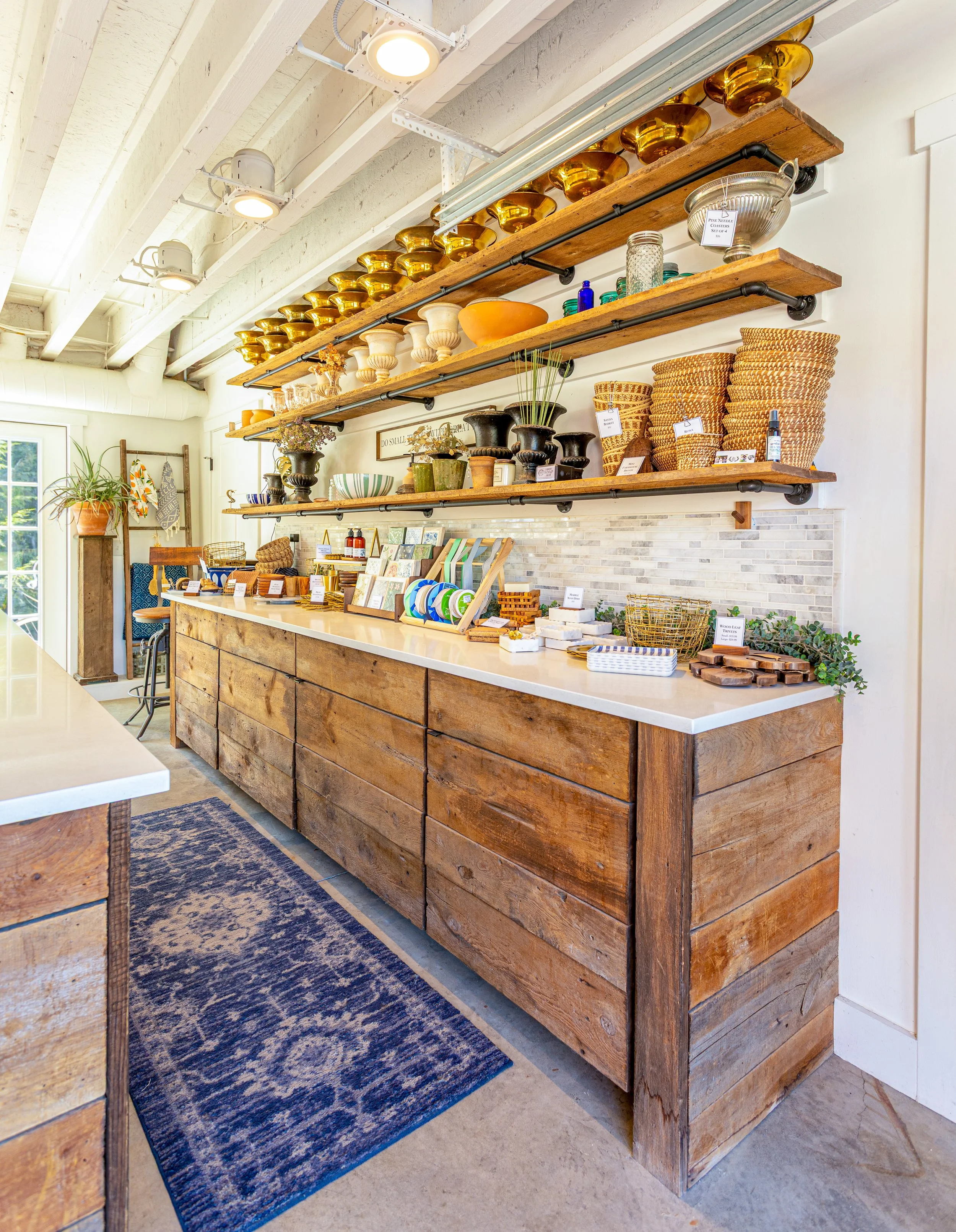 Interior of a store called Little Flower Soap Co., with wooden shelving displaying various decorative bowls, vases, and woven baskets, alongside a long wooden counter with small items and plants. A blue patterned rug lies on the concrete floor.