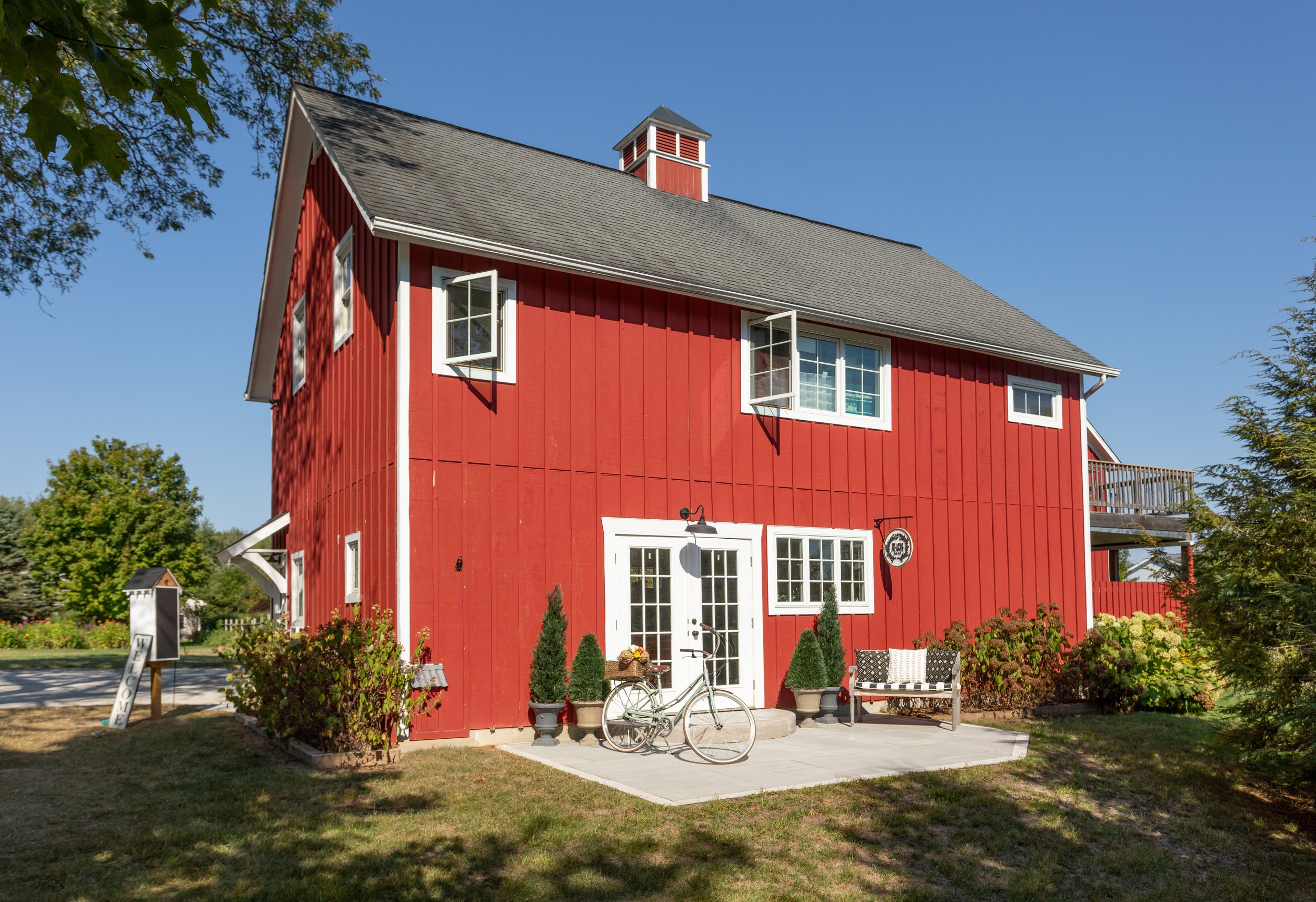 A two-story renovated barn for Little Flower Soap Co. with red paint and white trim. There is a white bicycle with a flower basket parked outside, with potted plants, a bench with pillows, and a welcome sign.