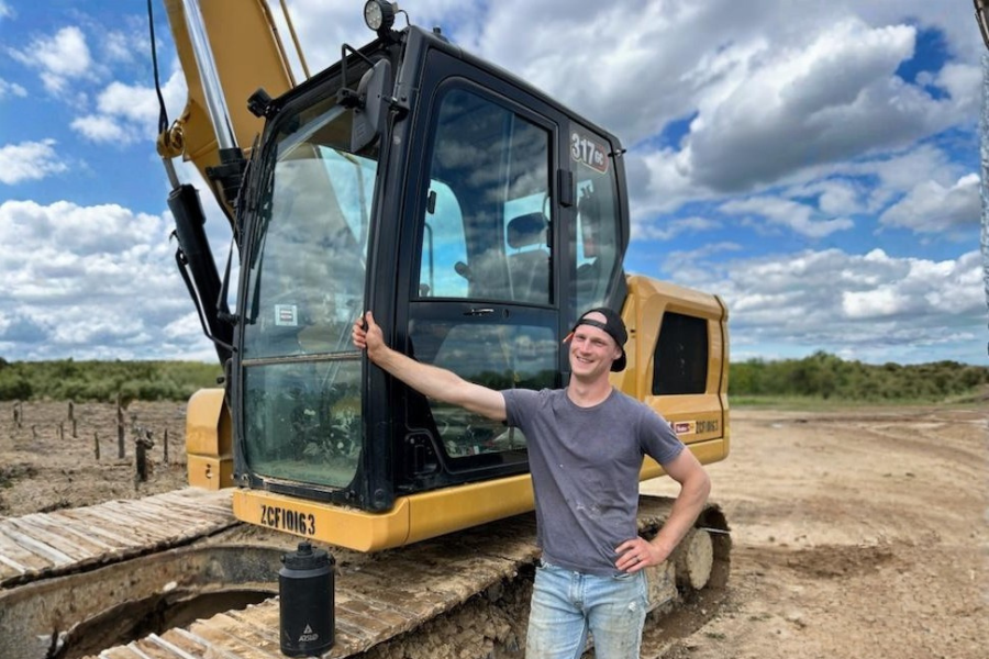 A smiling young man wearing a backward baseball cap and gray T-shirt standing next to a yellow excavator on a dirt construction site against a partly cloudy sky.