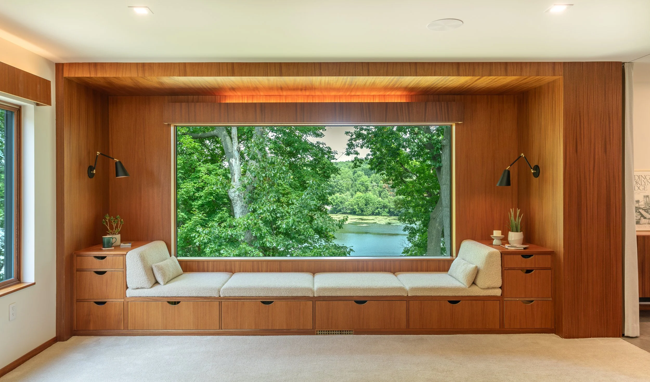 A built-in window seat reading nook in a mid-century modern home with custom renovations, with cushion and pillows, surrounded by wooden paneling, overlooking a lush green landscape with trees and a body of water.