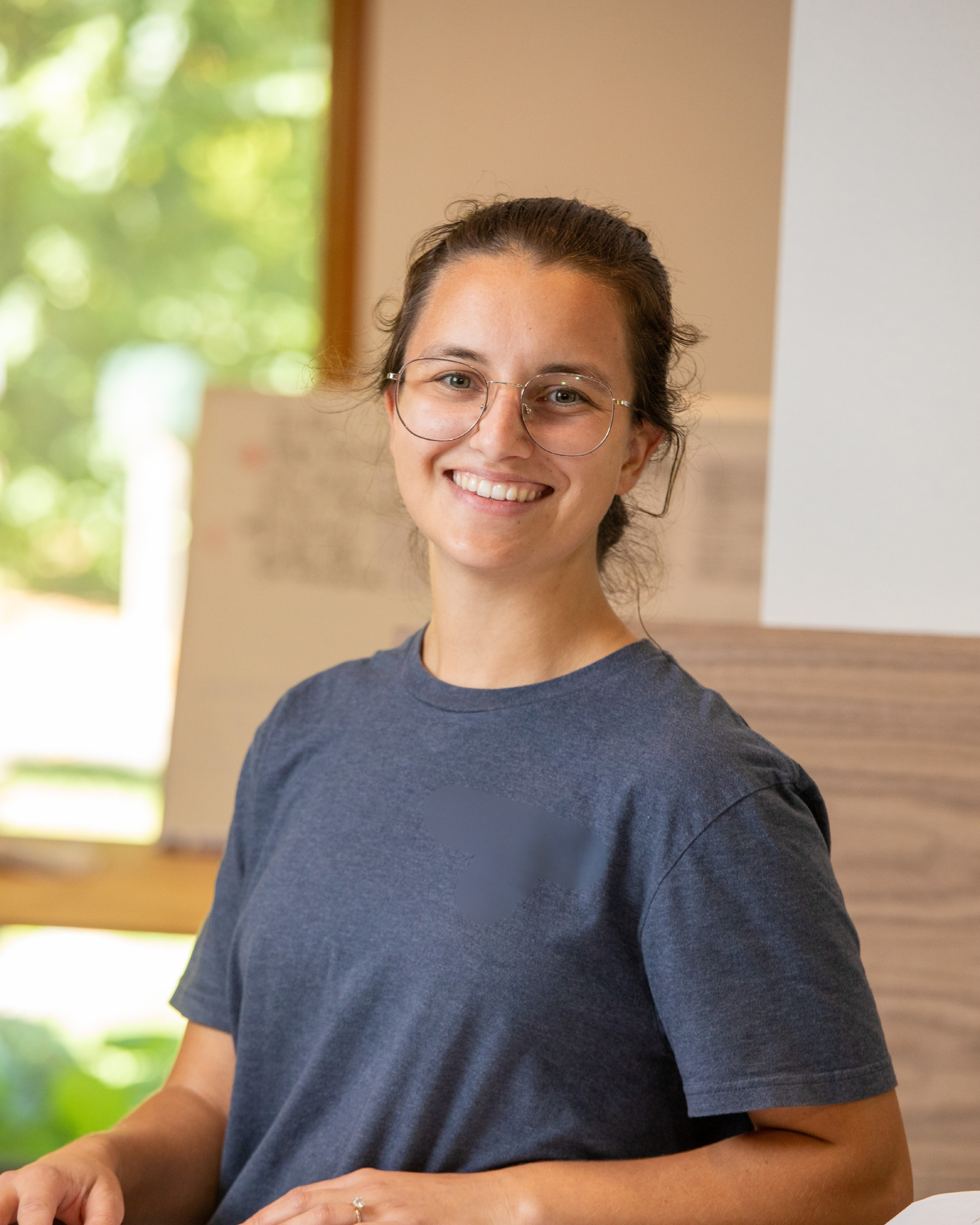 Rebecca Campbell is a smiling woman with glasses and brown hair tied back, wearing a dark gray T-shirt, standing indoors with a background of blurred windows and greenery.