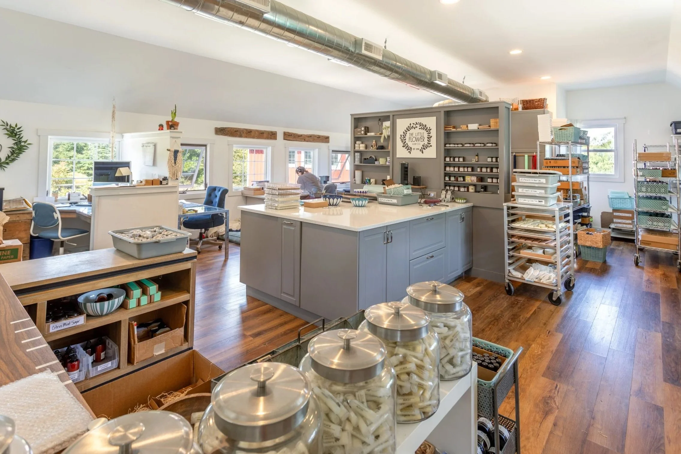 A bright and organized artisan shop called Little Flower Soap Co., with multiple windows, hardwood floors, and various shelving units holding supplies. There is a central island with bowls and supplies, with jars of products in the foreground.