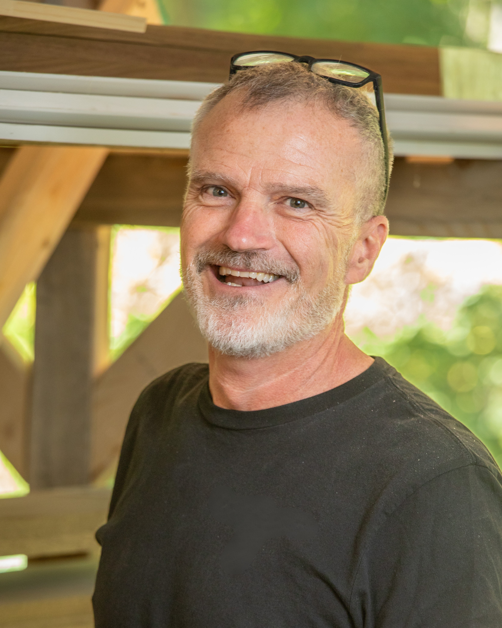 Steve Moody is a Lead Carpenter and a smiling middle-aged man with gray hair, beard, and glasses on his head, wearing a black shirt, outdoors with greenery and wooden structure in background.