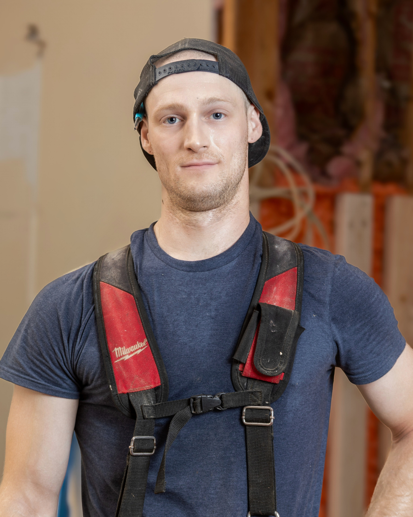 Robert Vandagriff is a young man wearing a backwards black baseball cap, navy blue t-shirt, and a black and red Milwaukee tool harness, standing in a construction or workshop setting with wooden beams and tools in the background.