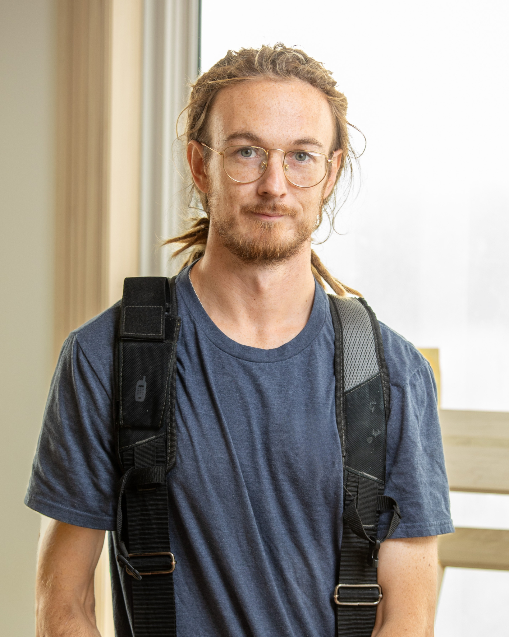 Josh Morris is a young man with glasses and long dreadlocks standing indoors near a window.