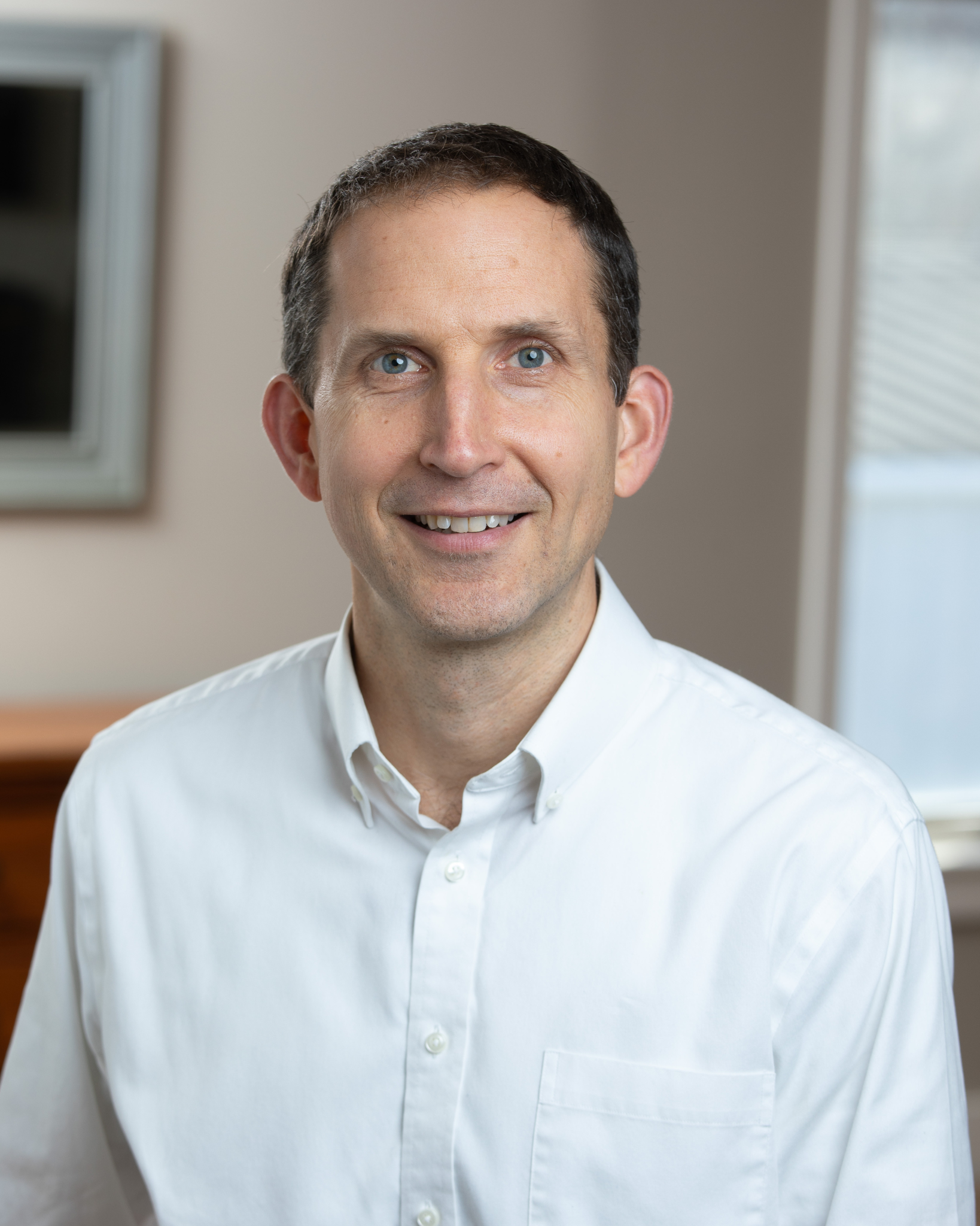 John Urbanic is a smiling middle-aged man with short dark hair wearing a white button-up shirt, indoors with a window and a blurred background.
