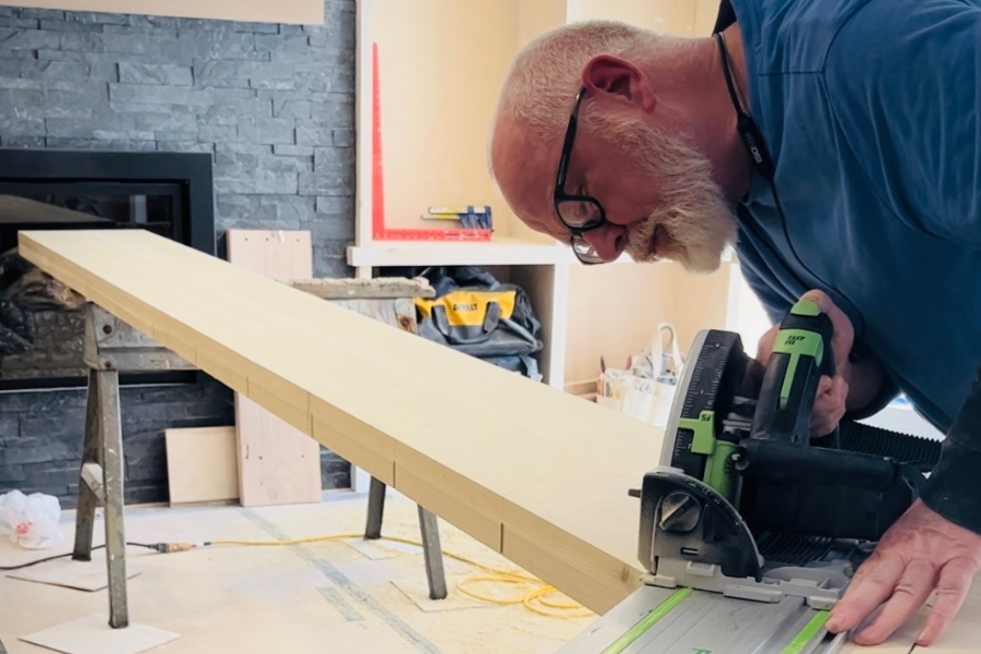 A man with a beard and glasses is cutting a long wooden board with a power saw in a workshop.