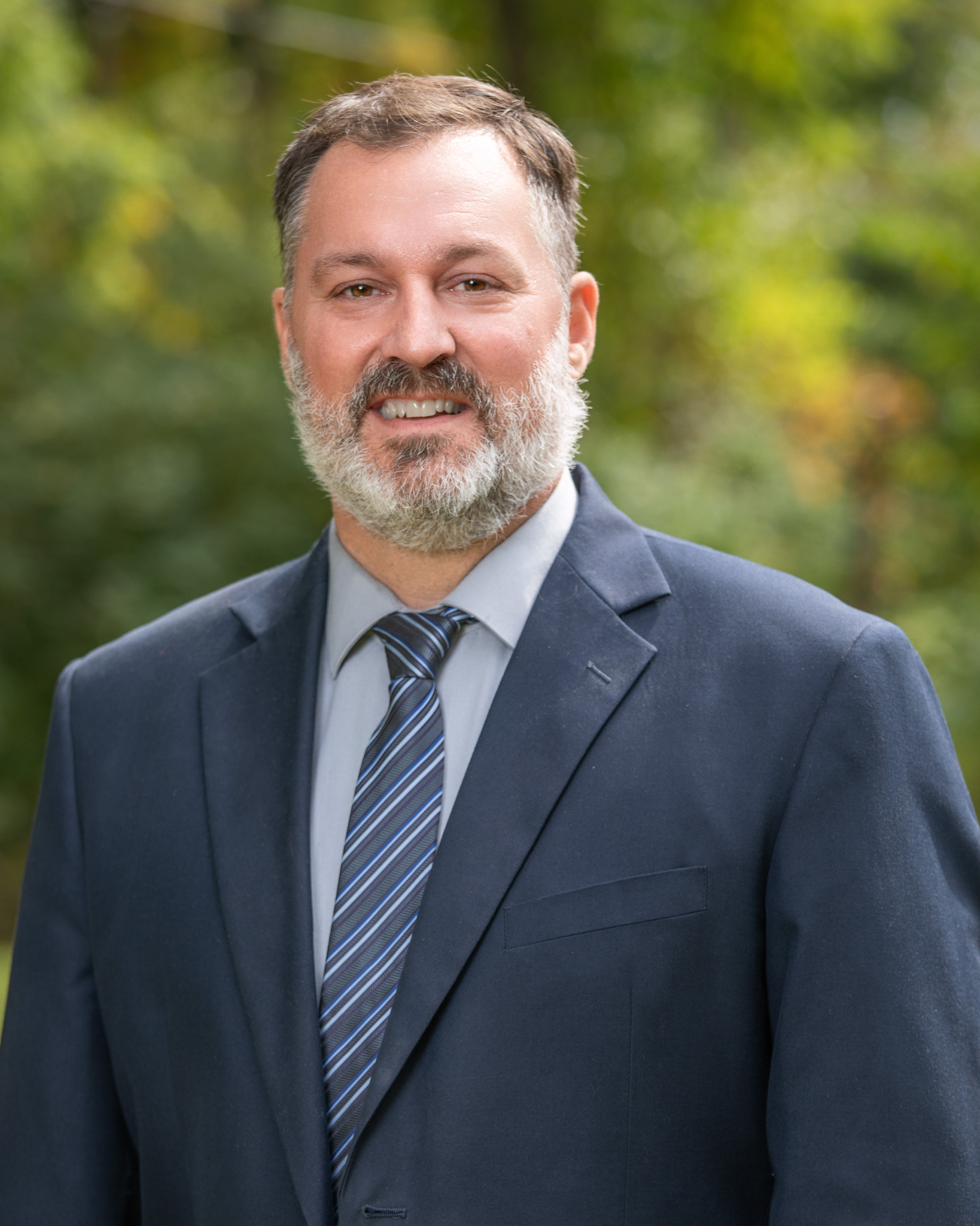 Mike Sumerton is a middle-aged man with a gray beard, wearing a dark blue suit, light blue shirt, and a striped tie, standing outdoors with a blurred green and yellow leafy background.