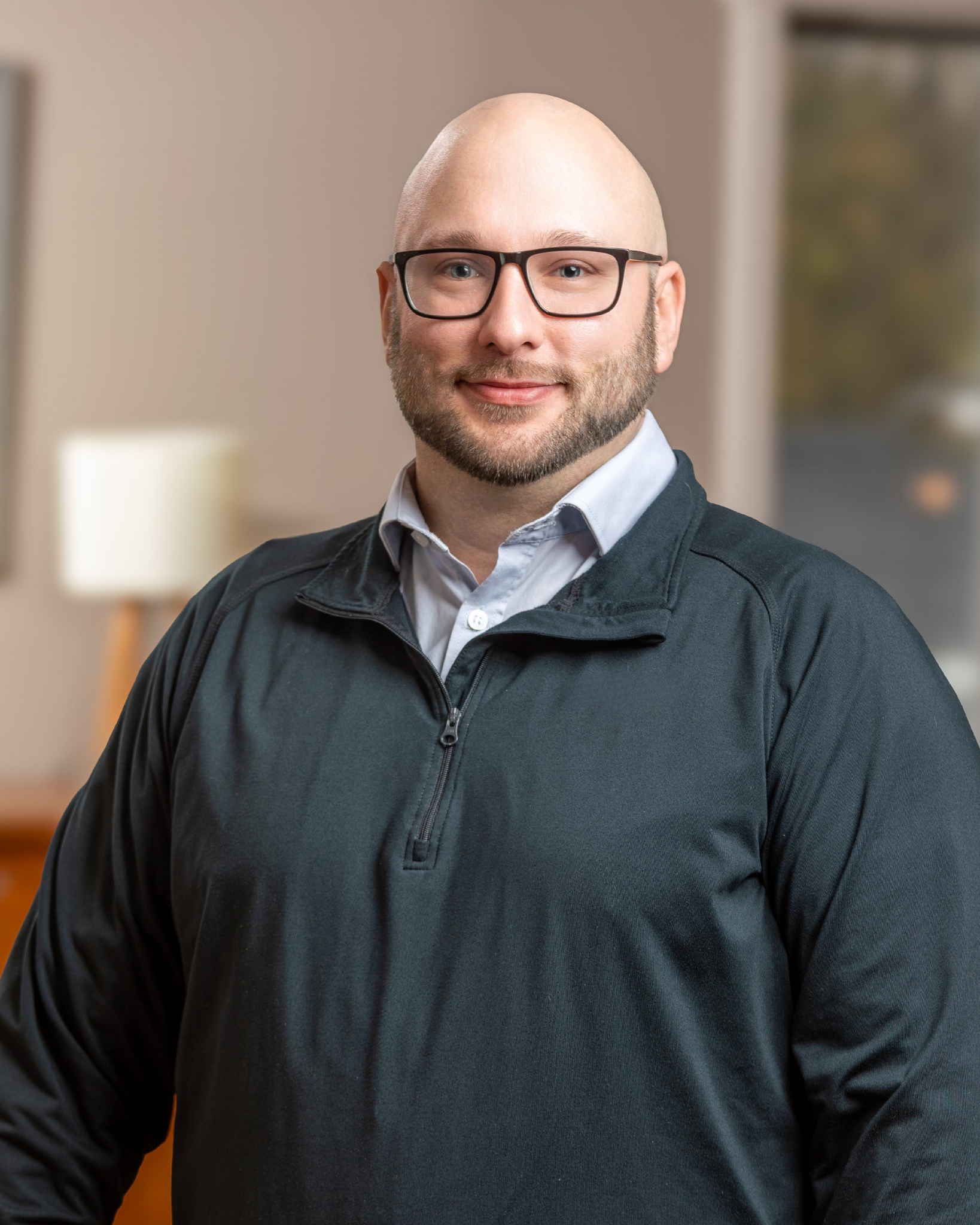 Bob Thomason is a middle-aged smiling man with glasses and a beard wearing a black jacket and white shirt, standing indoors with a blurred background.