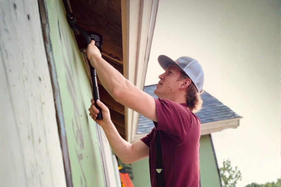 A young man using a power drill on the exterior of a house under construction.