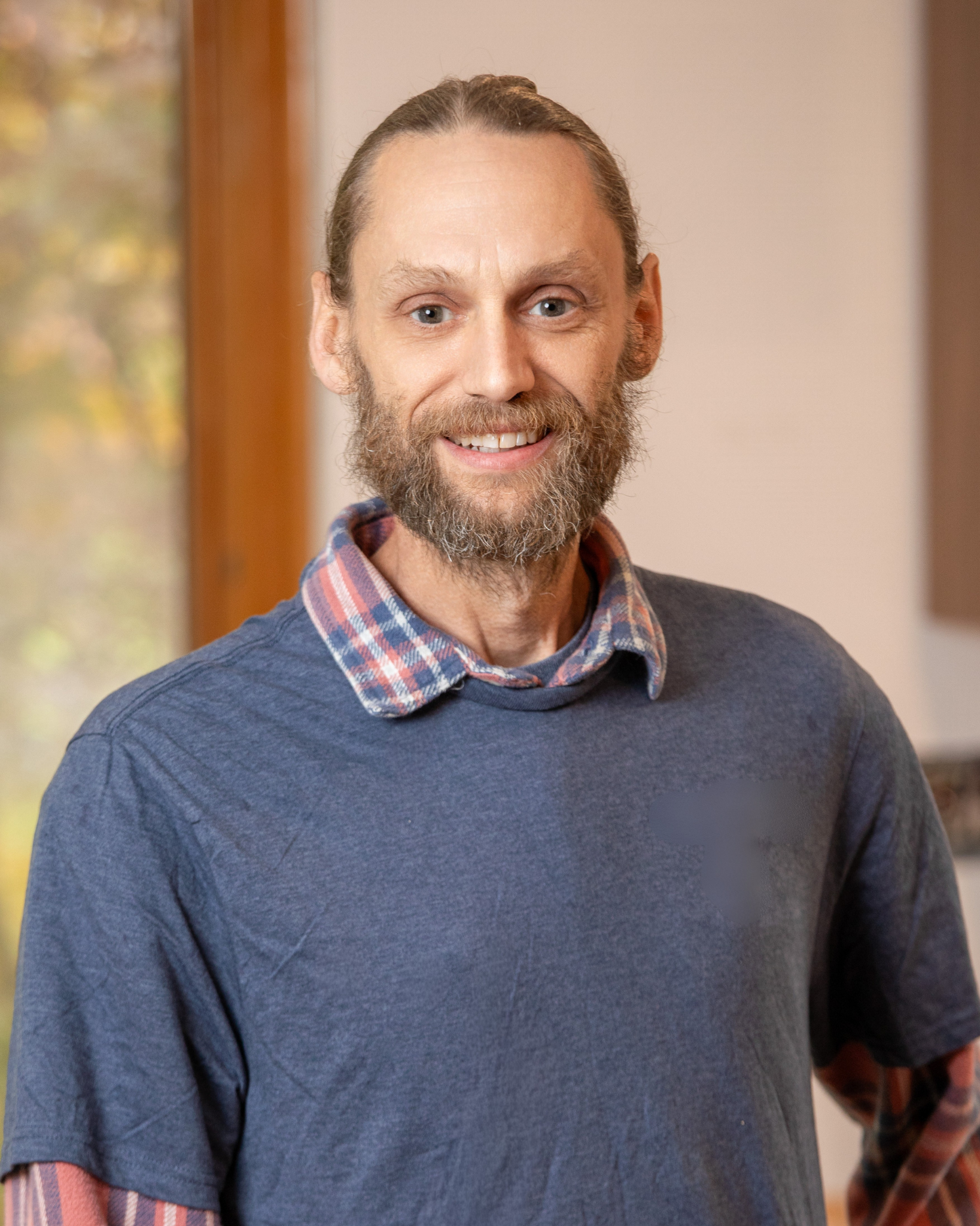 Keith Teacherworth is a middle-aged man with a beard, smiling, wearing a blue sweater over a plaid shirt, standing indoors near a window with trees visible outside.