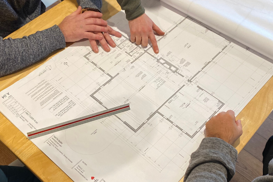Three people reviewing a large architectural floor plan spread out on a wooden table, with one person pointing at the plan and a metal scale ruler on the table.