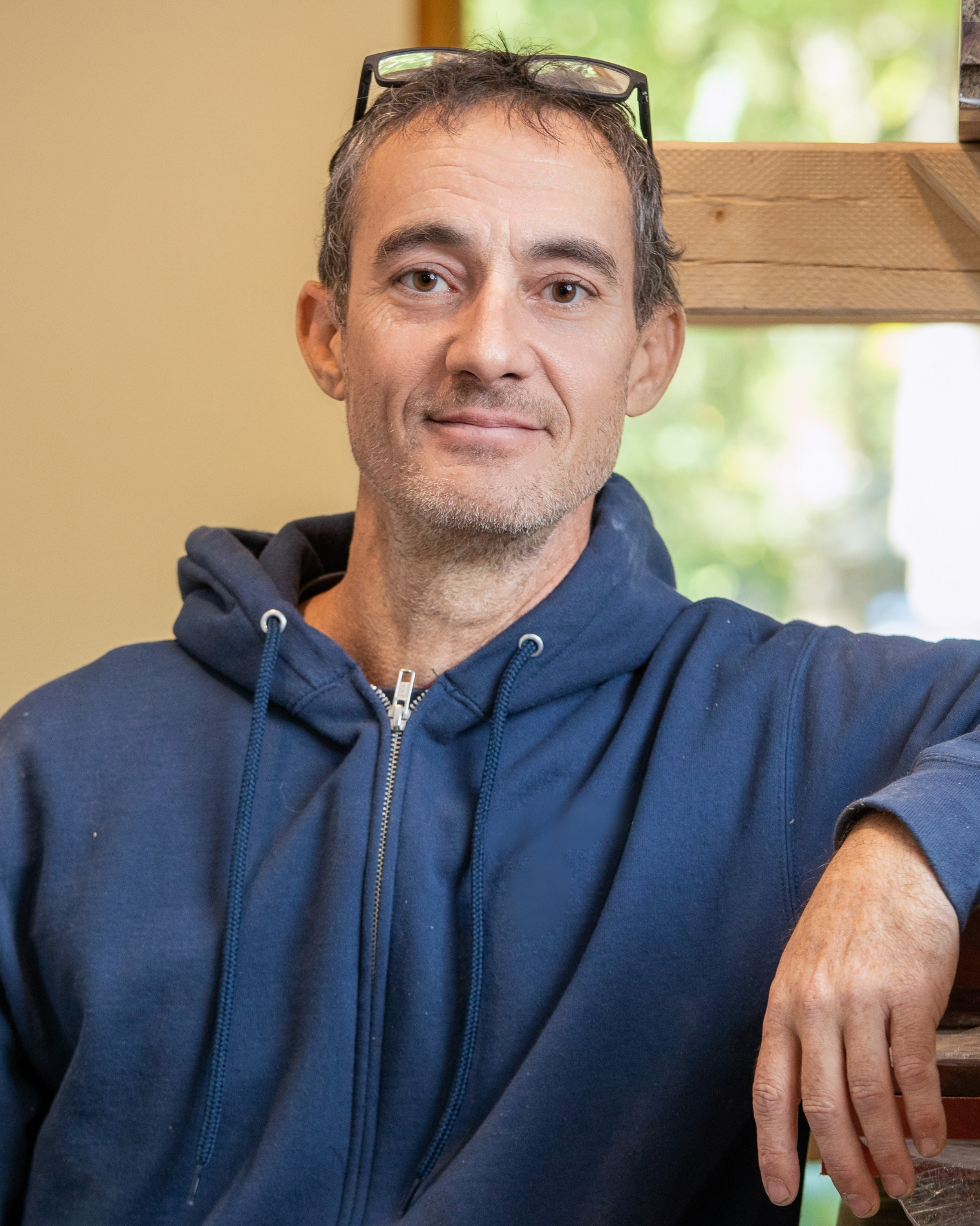 Kirk Gordon is a middle-aged man with glasses on his head and a blue hoodie, sitting indoors with a window behind him showing greenery outside.