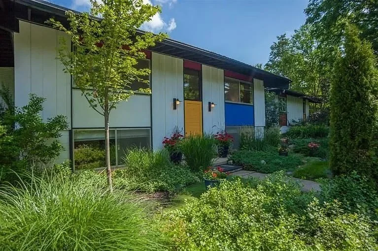 Modern house with a landscaped front yard featuring various green plants, trees, and colorful flowers, under a blue sky with some clouds. This home was originally designed by Edward Bartolf “Ted” Smith, Jr., a graduate of the University of Michigan.