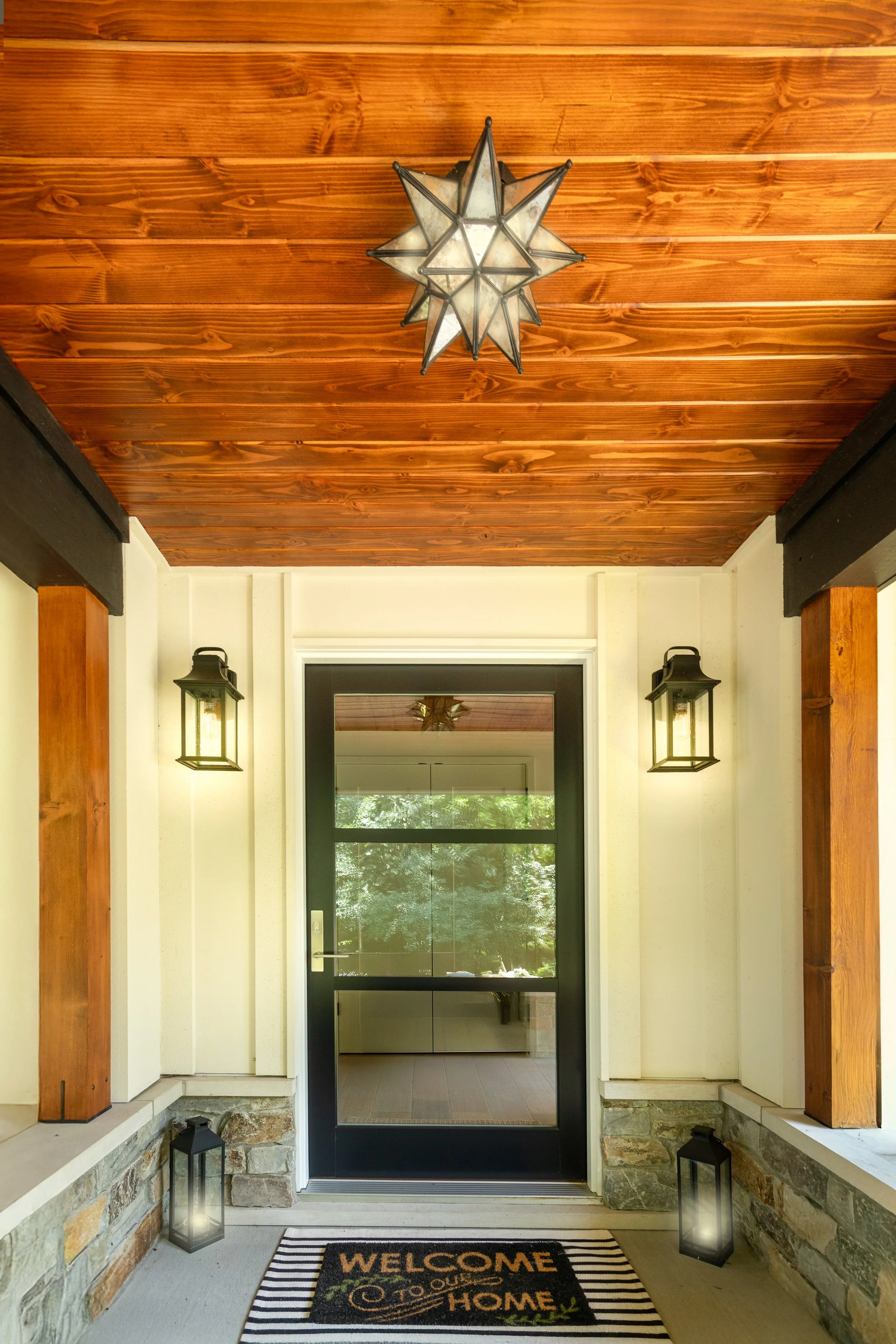 Front porch with black door, stone accents, striped welcome mat, wall-mounted lanterns, and a star-shaped ceiling light.