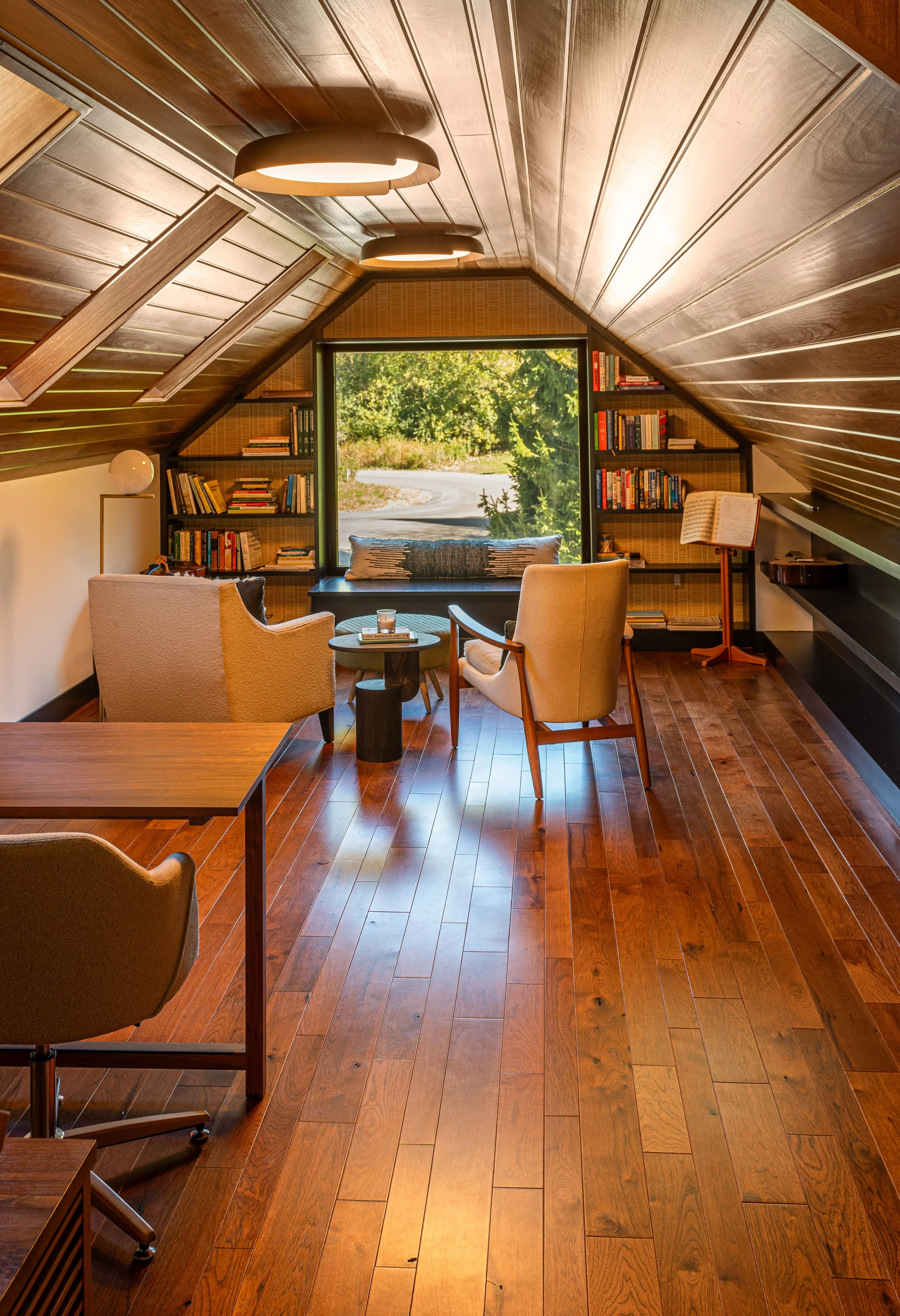 Cozy attic reading nook with wooden ceiling, bookshelves, a window seat, two armchairs, and a view of greenery outside.