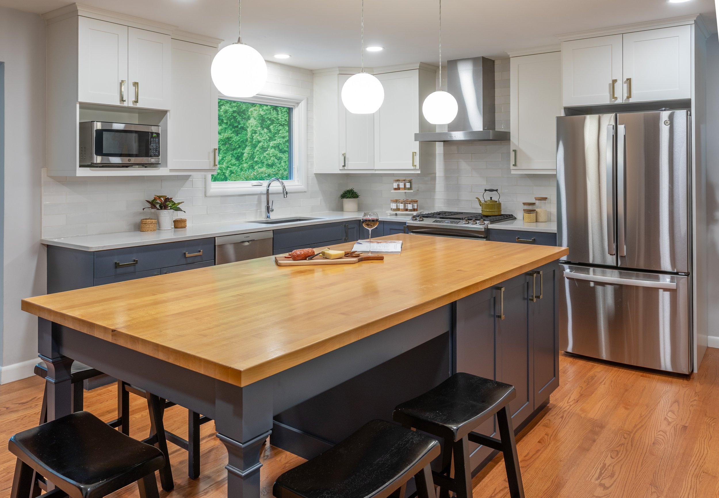 Modern kitchen with white upper cabinets, dark lower cabinets, stainless steel refrigerator, microwave, and oven, a large wooden island, and three pendant globe lights, with a window showing greenery outside.