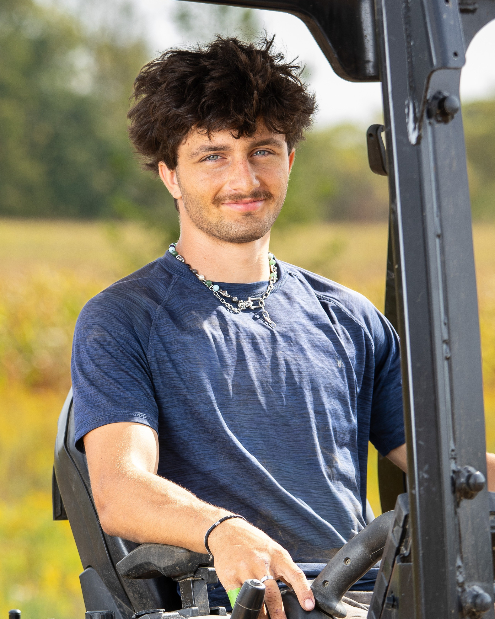 Vincent Redente is a young man with tousled dark hair, blue eyes, and a slight smile, sitting in a bulldozer in an outdoor setting during daytime, wearing a blue T-shirt, a beaded necklace, and a black wristband.