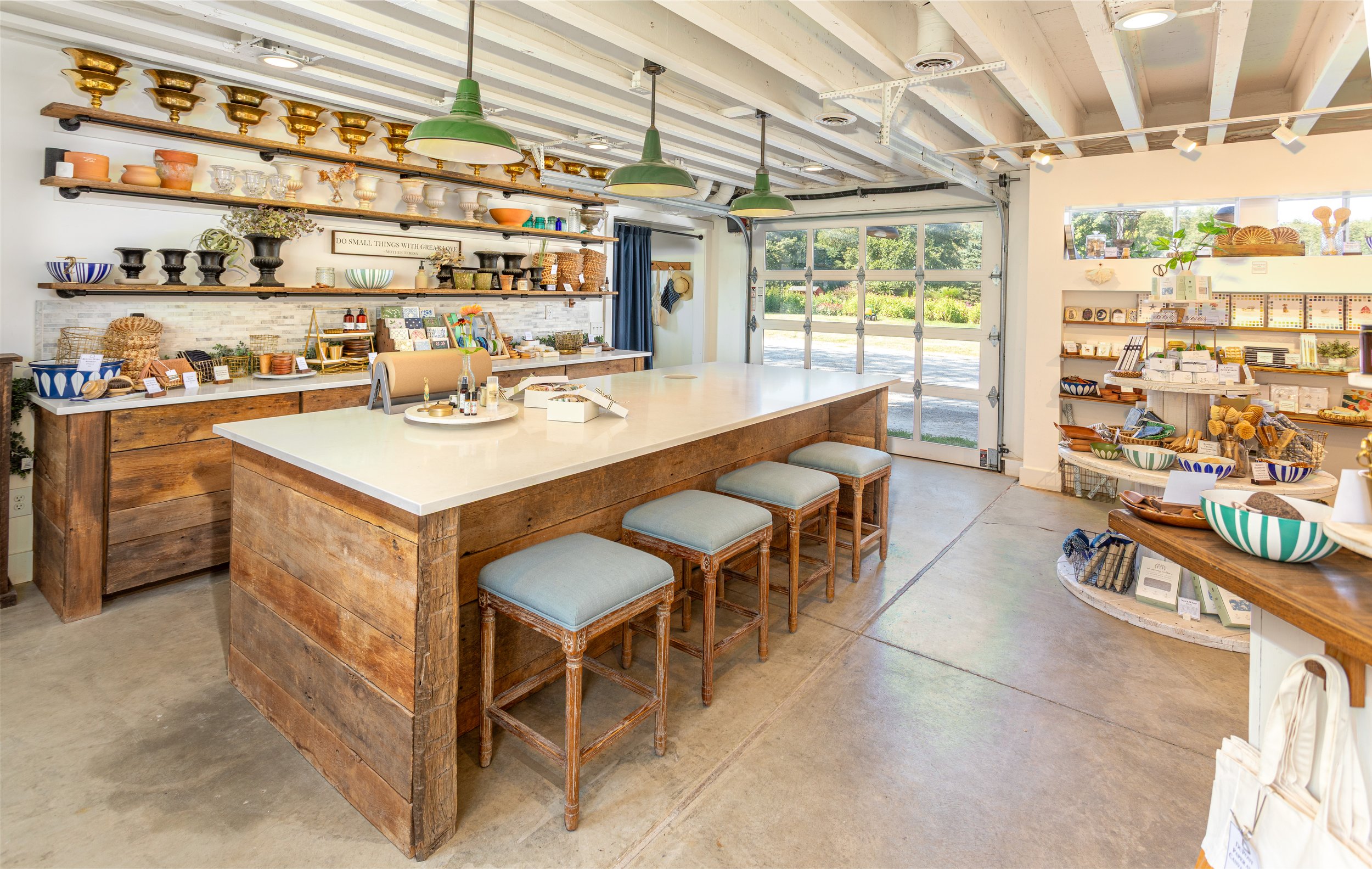 Interior of a retail shop selling hand-made artisan soaps, featuring wooden furniture, open shelves with various bowls, vases, and decorative items, a large island with stools, and large windows letting in natural light.