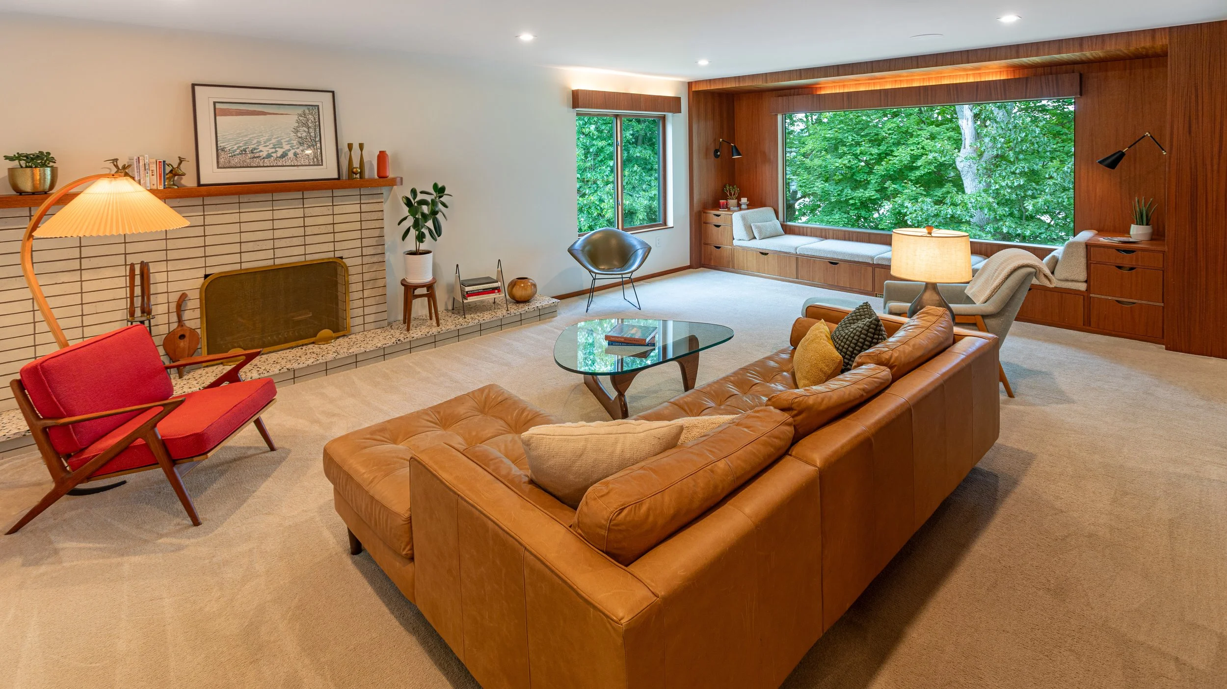 Spacious living room with large window view of green trees, tan leather sectional sofa, glass coffee table, red mid-century lounge chair, beige armchair, fireplace with decorative items, and a built-in window seat.