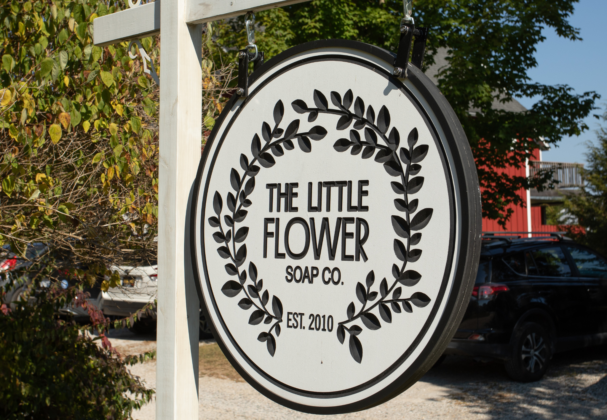 Sign for The Little Flower Soap Co., established in 2010, hanging outdoors with trees and parked cars in the background.