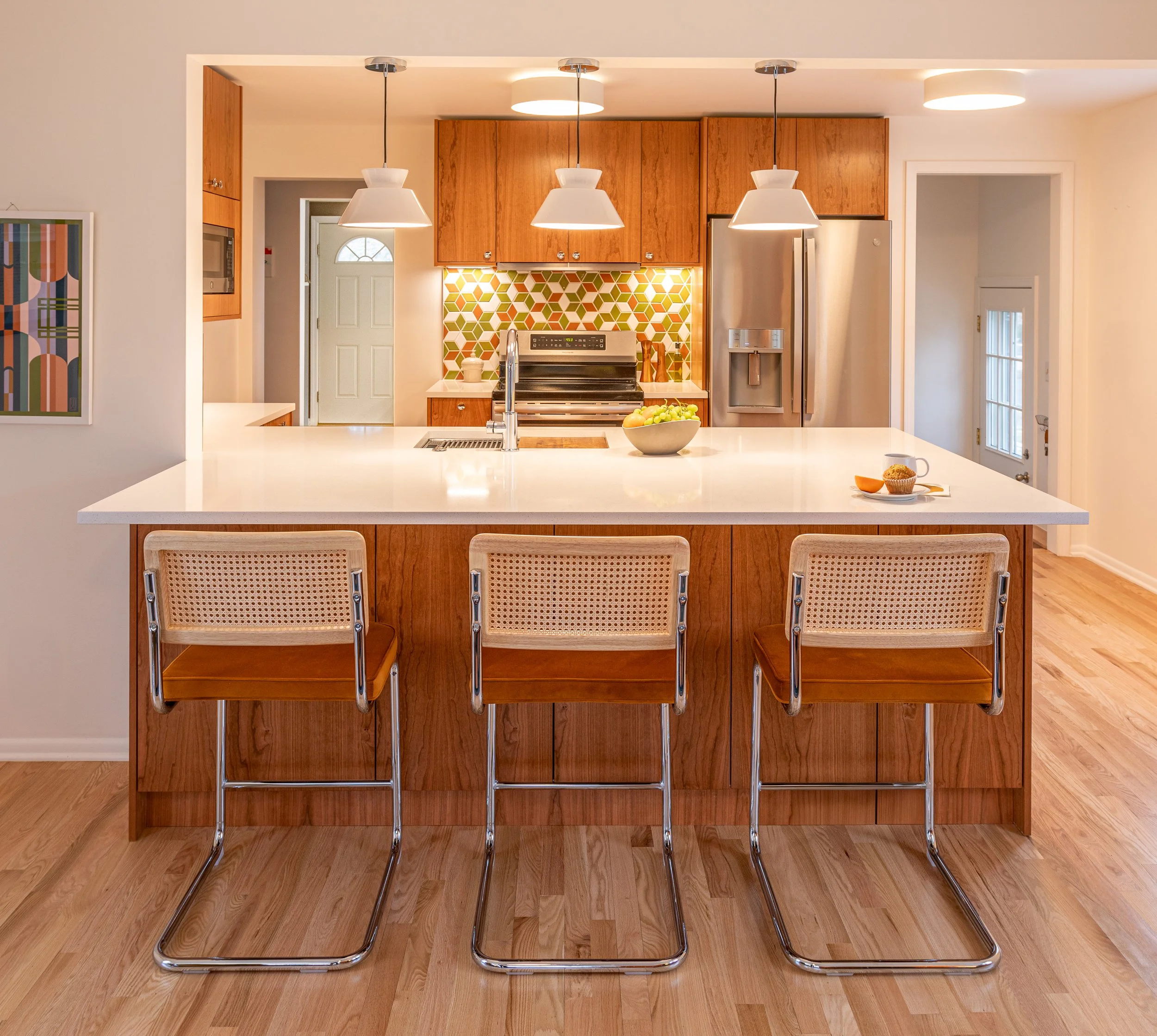 Modern kitchen with wooden cabinets, white island, colorful green and orange tile custom backsplash, stainless steel refrigerator, and three bar stools.