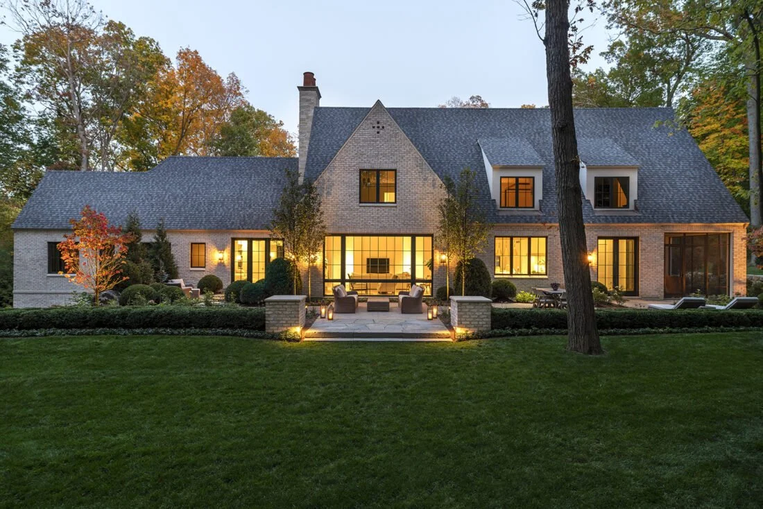 A modern two-story house with large windows, surrounded by greenery, illuminated at dusk with outdoor seating and landscaping.