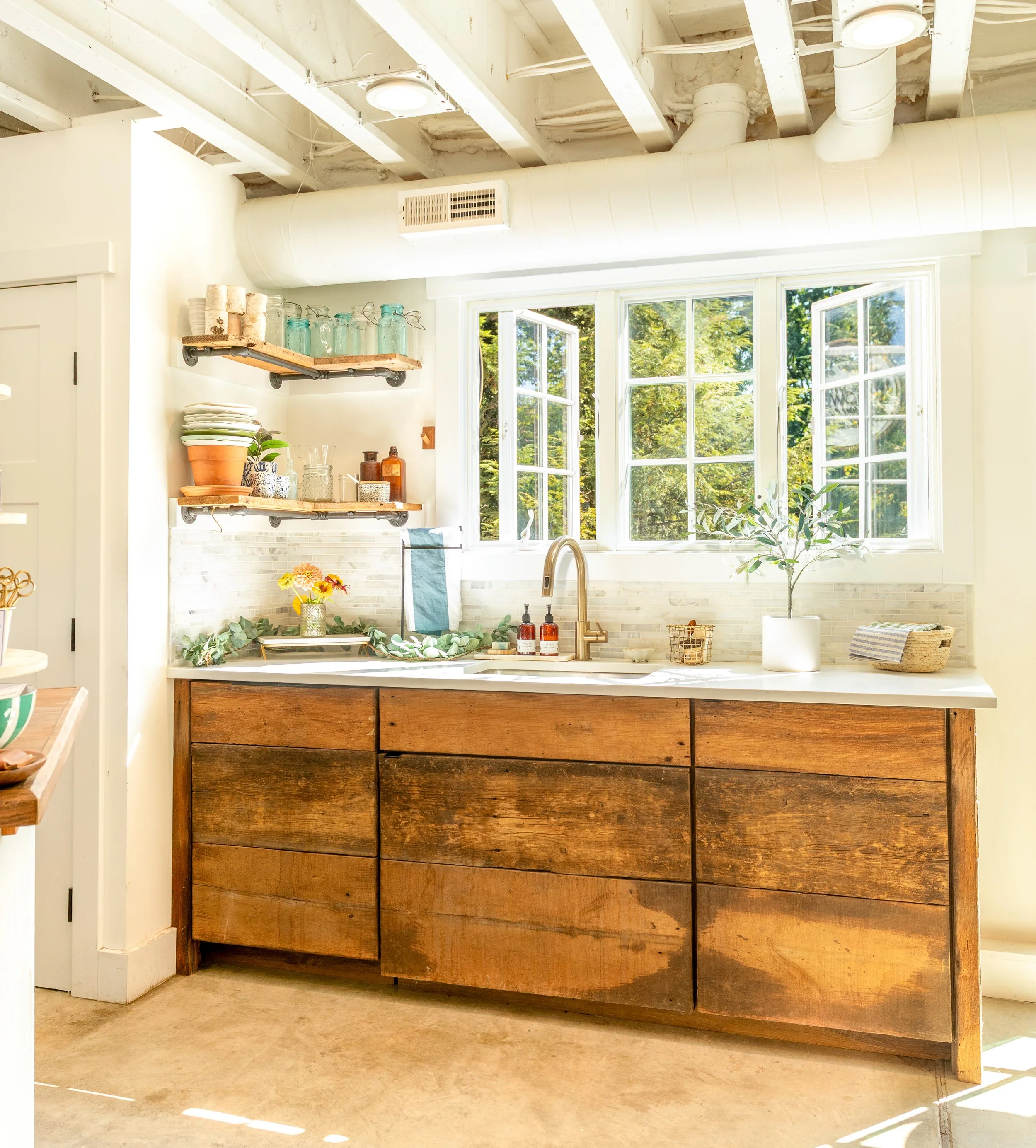 Bright kitchen work space at Little Flower Soap Co., with large window, wooden cabinets, potted plant, open shelves with jars and dishes, and a stainless steel sink.