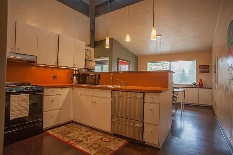 Kitchen with white cabinets, black stove, orange backsplash, wooden countertop, hanging pendant lights, and a window letting in natural light. This is a "Before" photo that shows a client's home before we renovated it.